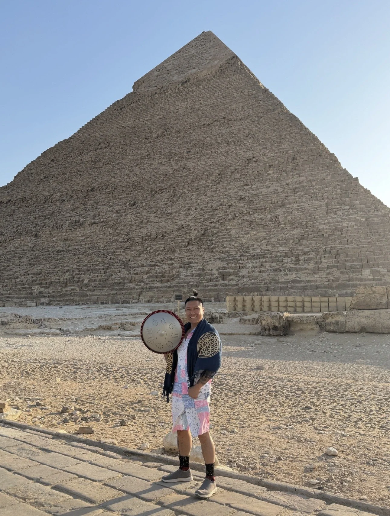 Alex Ma standing in front of the Great Pyramid of Giza, holding a steel tongue instrument, wearing casual clothing and smiling.