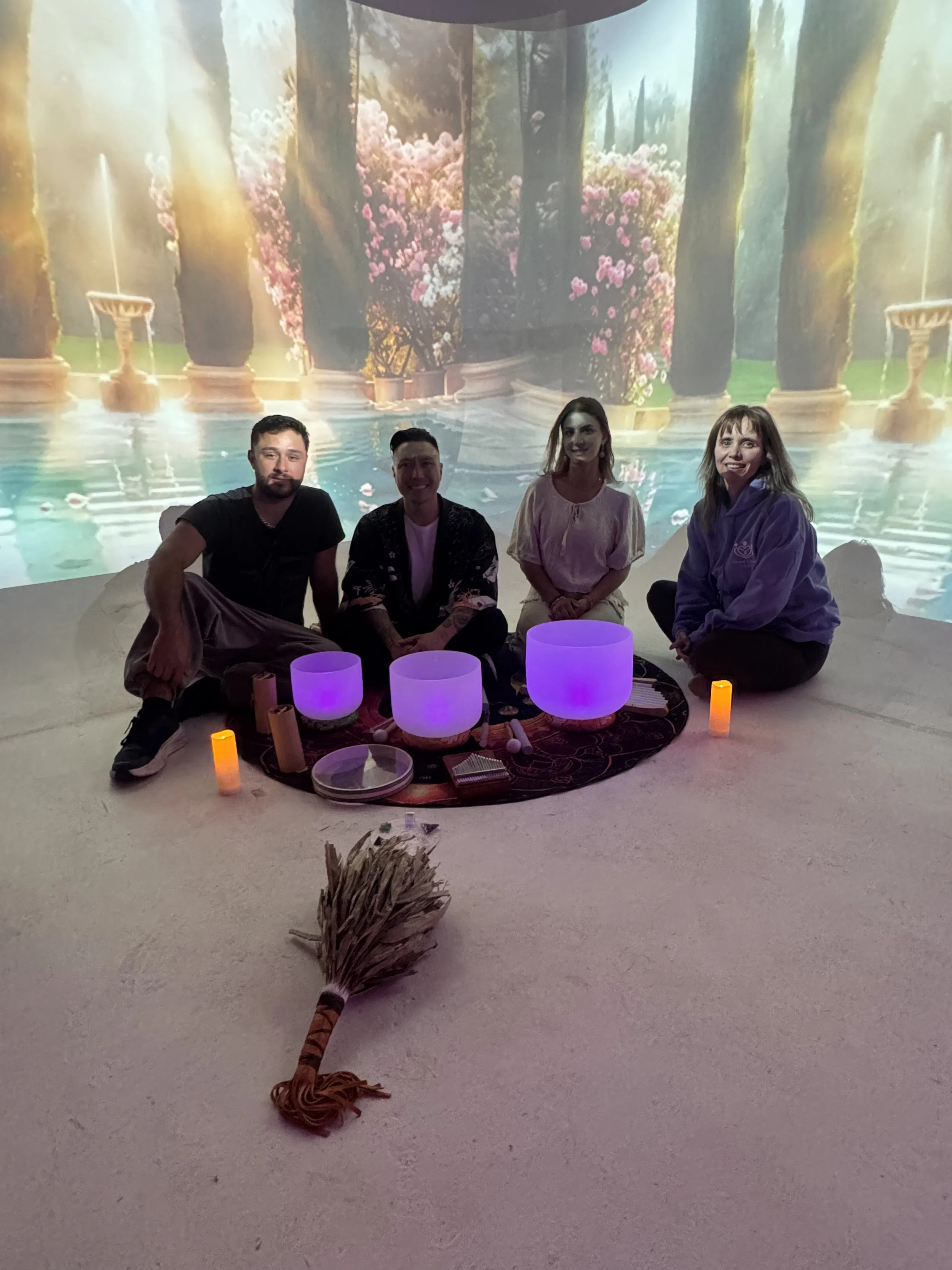 Alex Ma, Samper Caicedo, and Saja Light sitting on the floor in front of a scenic, indoor projection of a garden with columns and pink flowers. They are surrounded by candles and a mat with singing bowls, a drum, and a chakapa. After a sound bath per