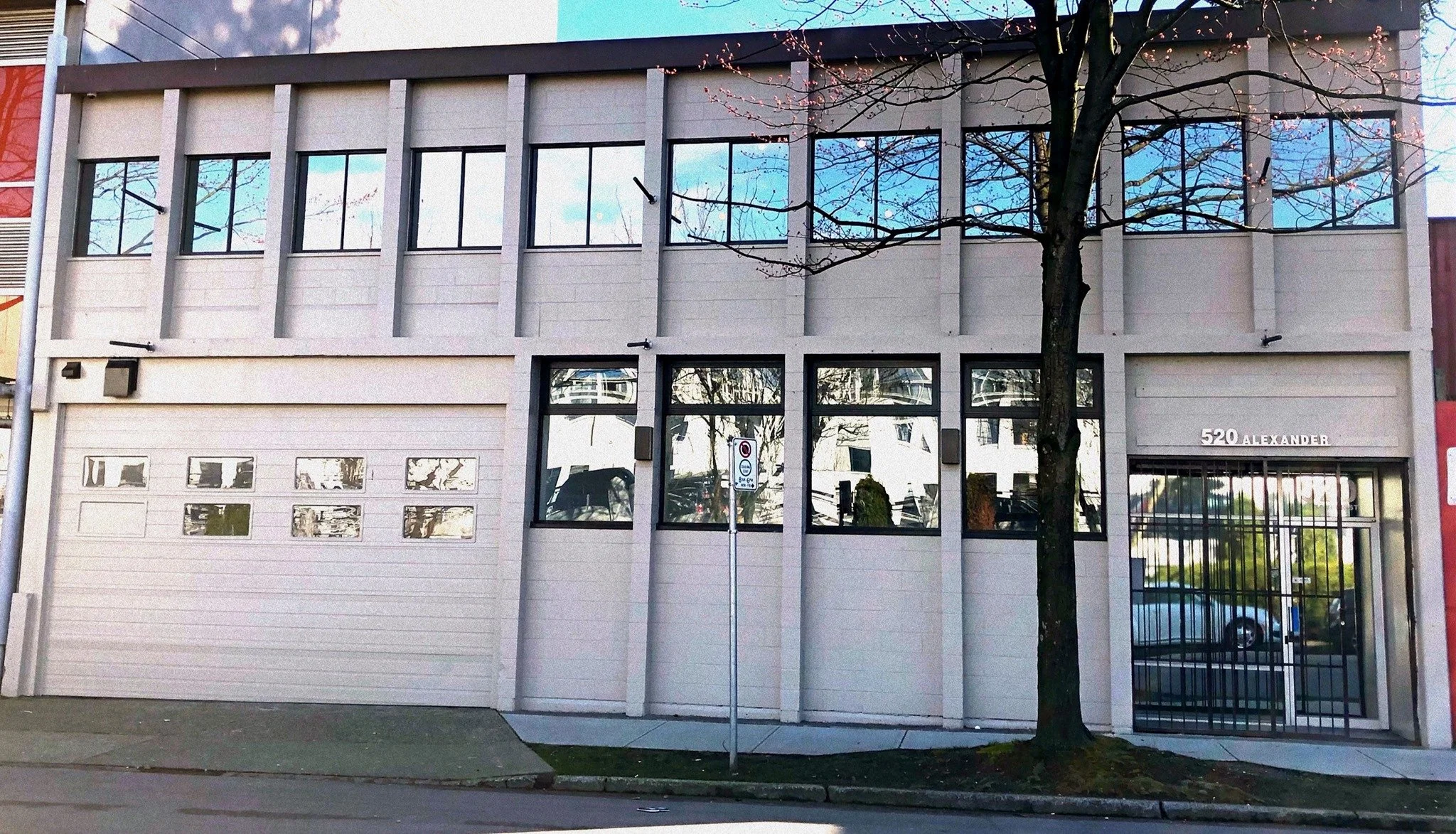 520 Alexander St venue. A modern two-story building with a gray facade and large windows on both floors reflecting the sky and surrounding trees. There is a tree in front with pink blossoms, and a street sign and parked cars are visible nearby.