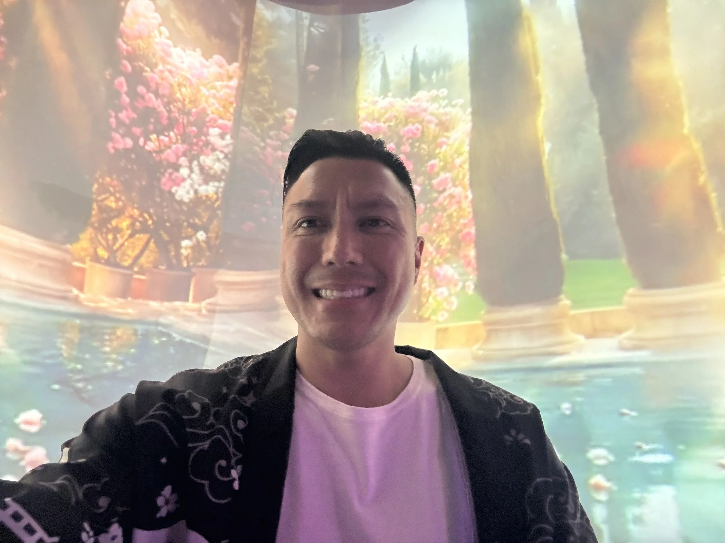 Alex Ma from Solar Soul Circle taking a selfie in front of a colorful nature-themed digital display or mural featuring trees, pink flowers, and a pond. For his sound bath in Gastown, Vancouver, BC.
