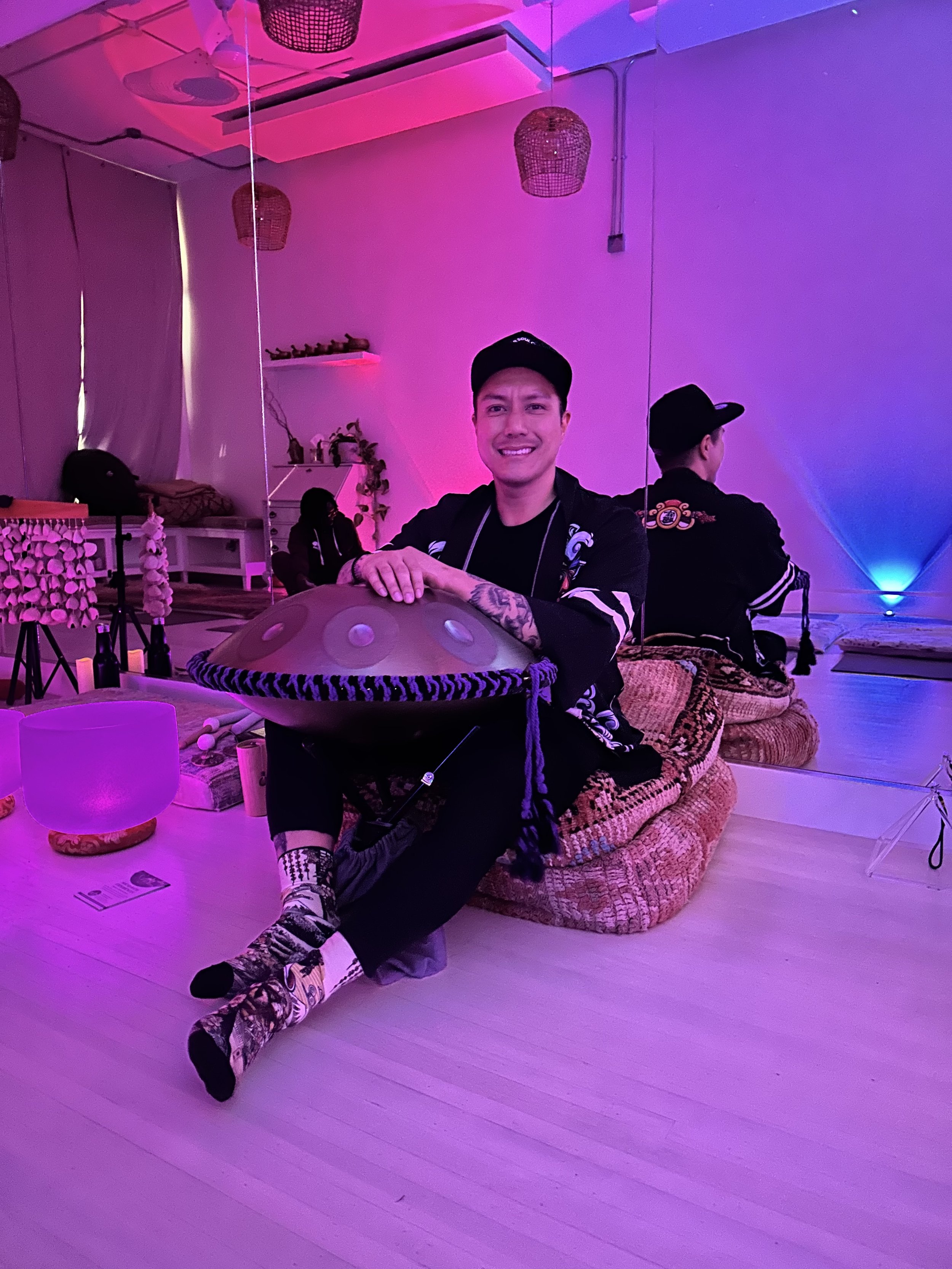 Alex ma from Solar Soul Circle in a black cap and clothing with patterned socks sits on a colorful woven blanket on the floor, leaning on a large handpan instrument in a room bathed in pink and purple lighting. Behind him, a mirror reflects his back 