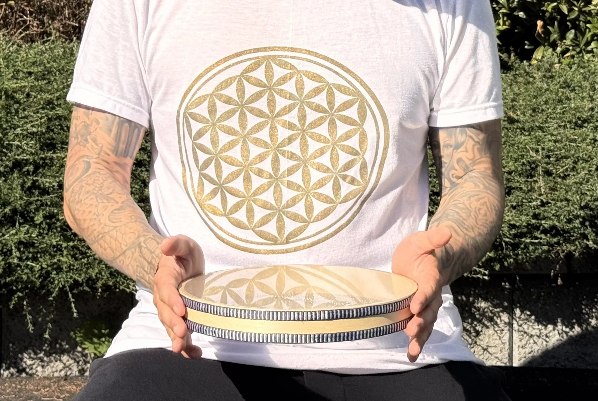 Alex Ma from Solar Soul Circle playing the ocean drum outdoors on a sunny day. Wearing a white flower of life shirt.