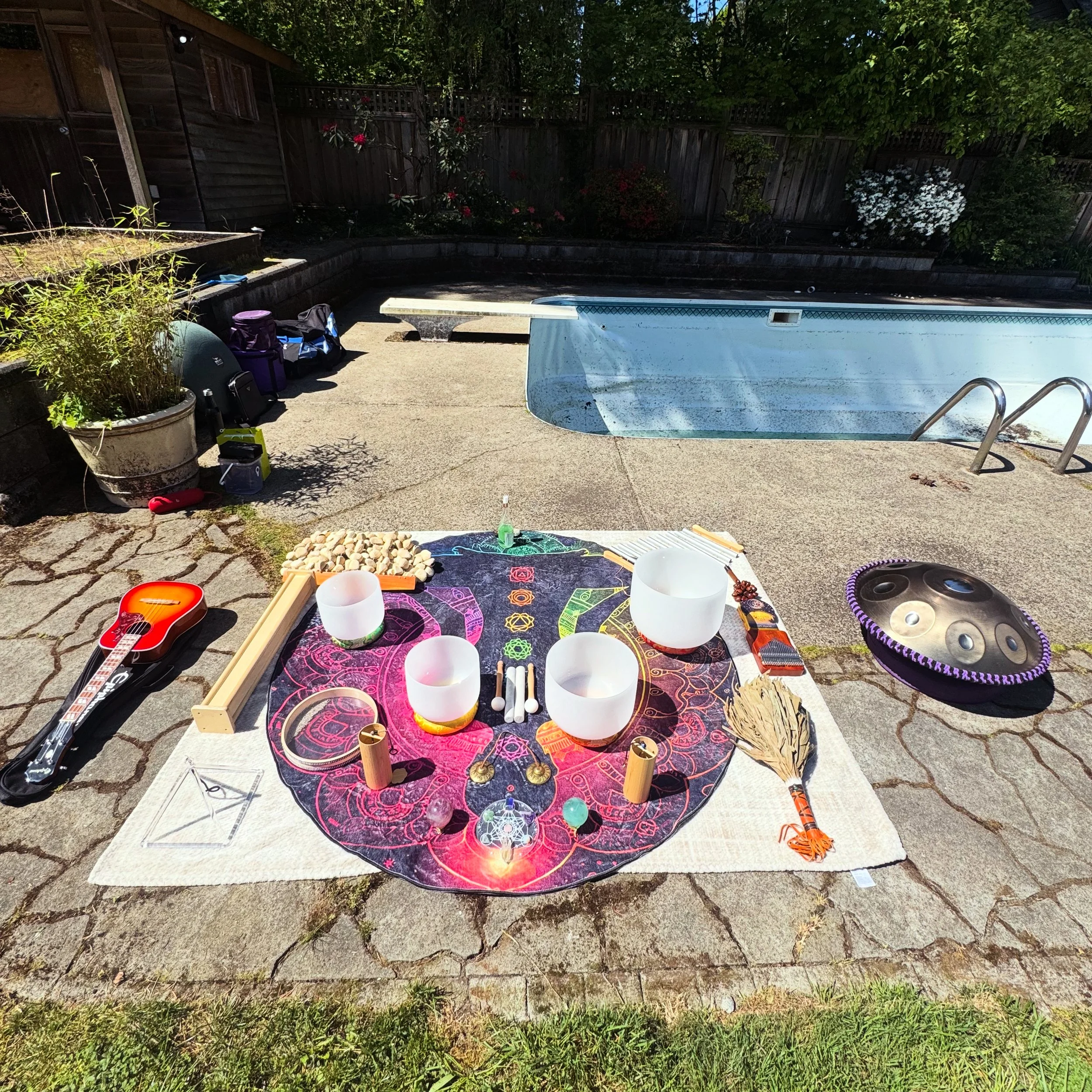 Backyard birthday sound bath photo with empty swimming pool, various musical instruments and objects placed on a large colorful tapestry, set against a green fence and trees. Located in North Vancouver BC.