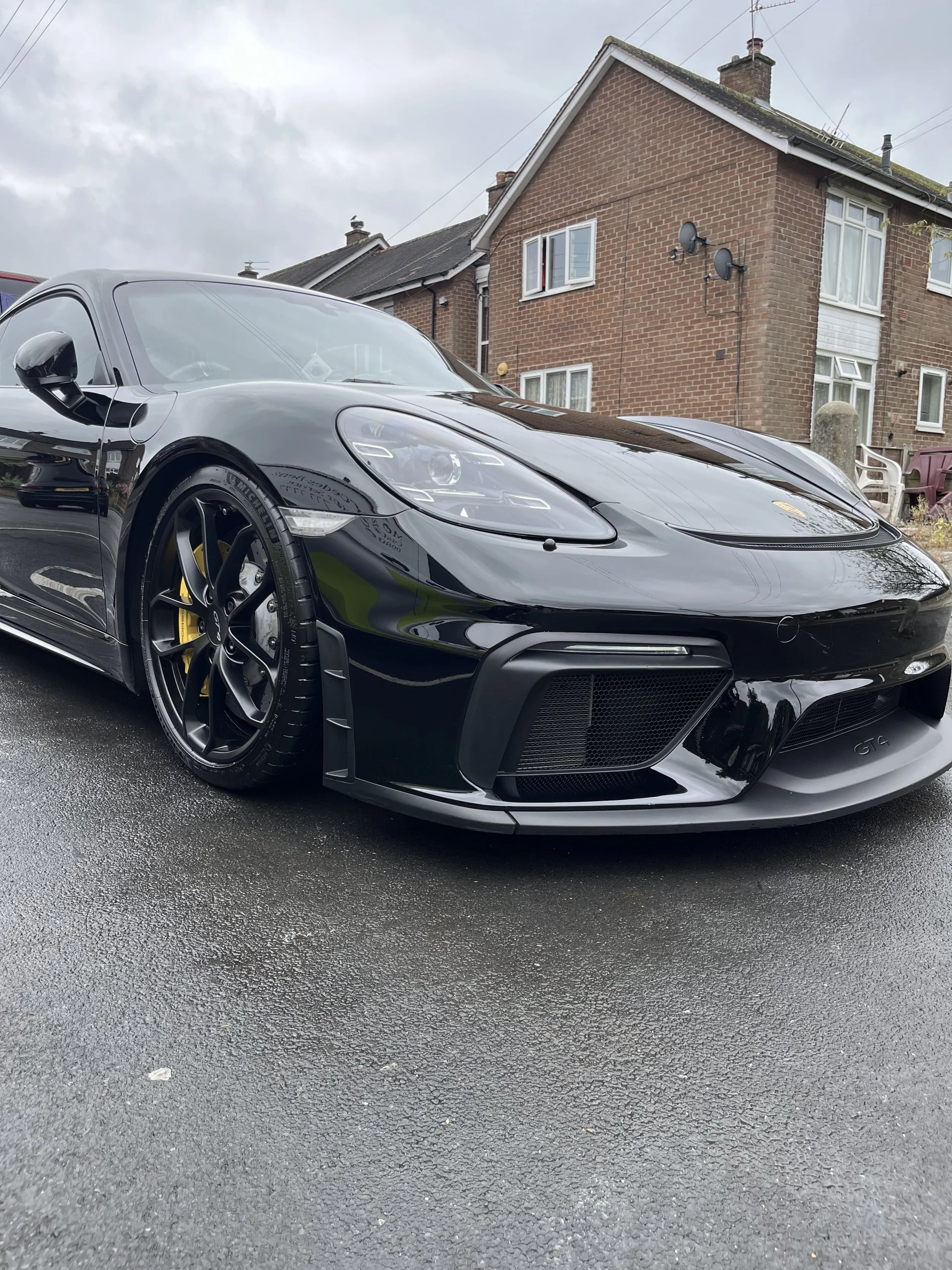 A black sports car parked on a wet driveway in front of a brick house with white window frames, cloudy sky in the background.
