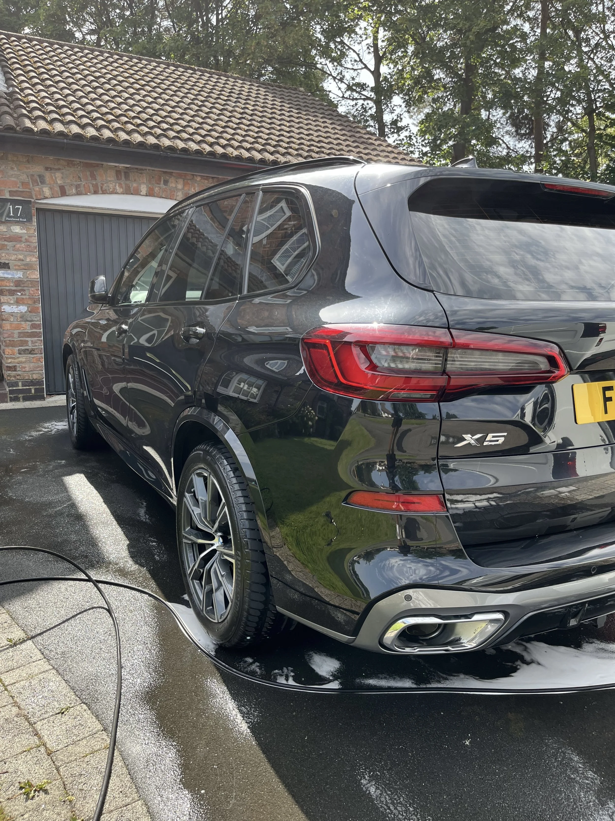 Black BMW X5 SUV being washed, with soap suds on the ground and water on the car, parked outside a house with brick walls and a tiled roof.