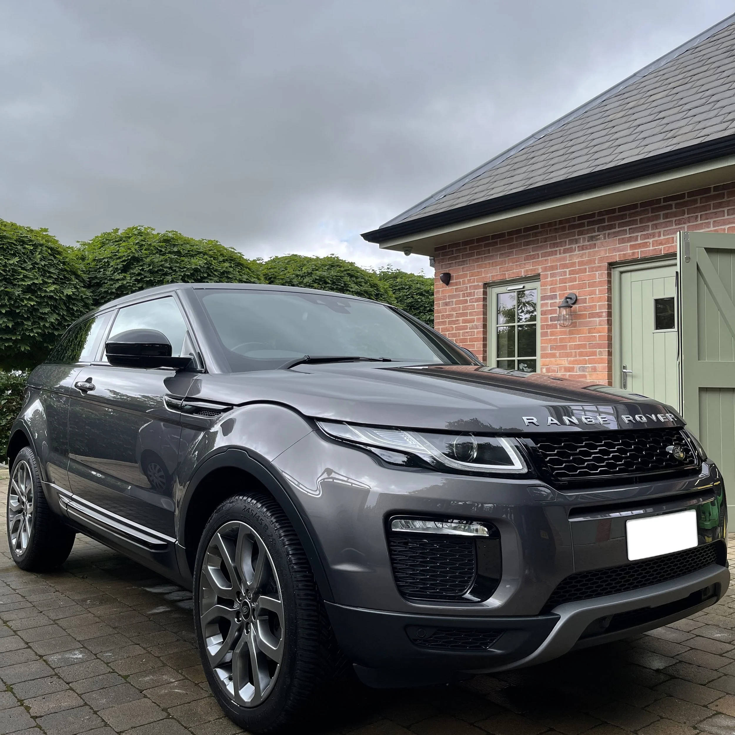 Gray Range Rover parked in front of a brick house with green doors and cloudy sky.