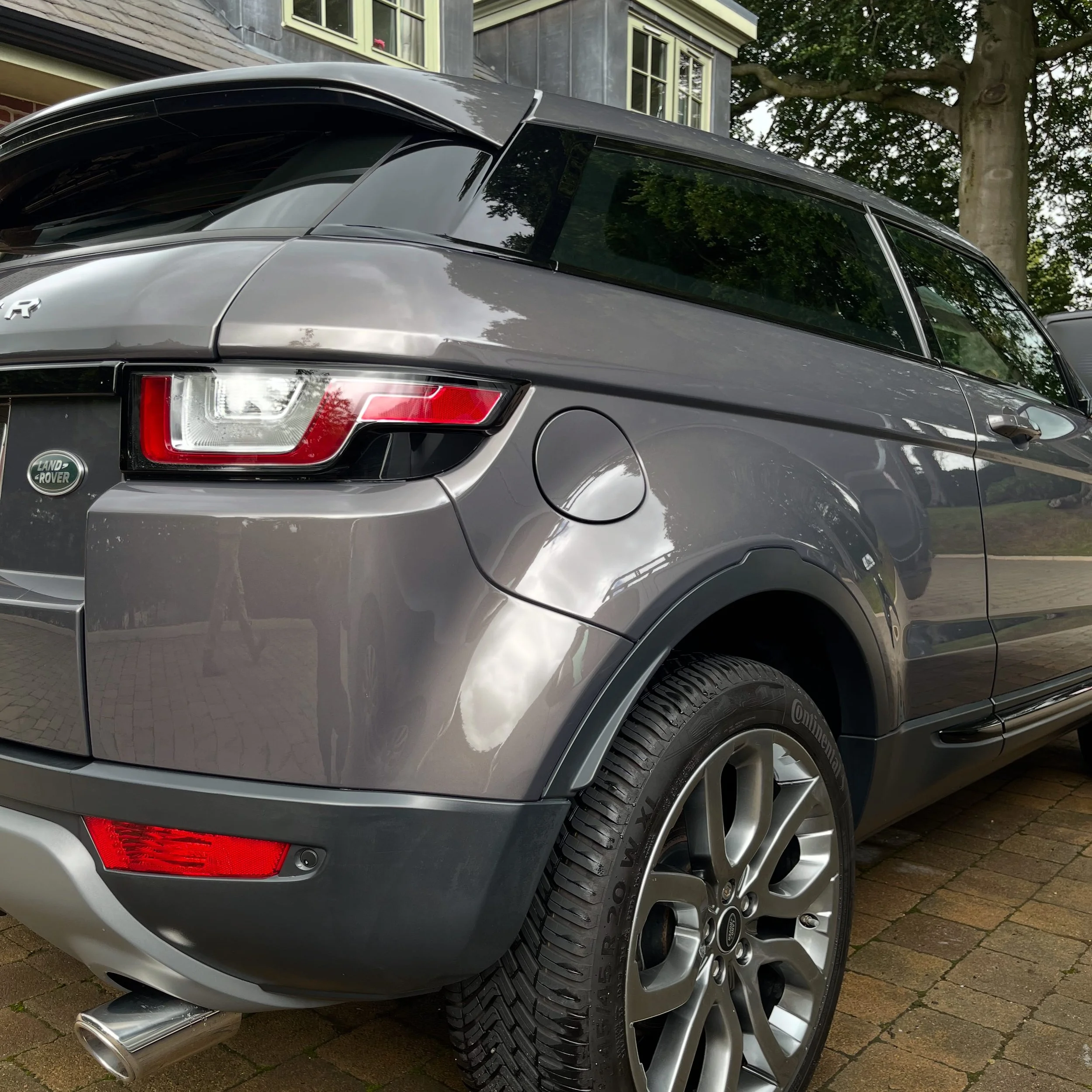 Close-up of the rear side of a gray Land Rover SUV parked on a brick driveway, showing the tail light, wheel, and exhaust pipe, with a garden and house in the background.