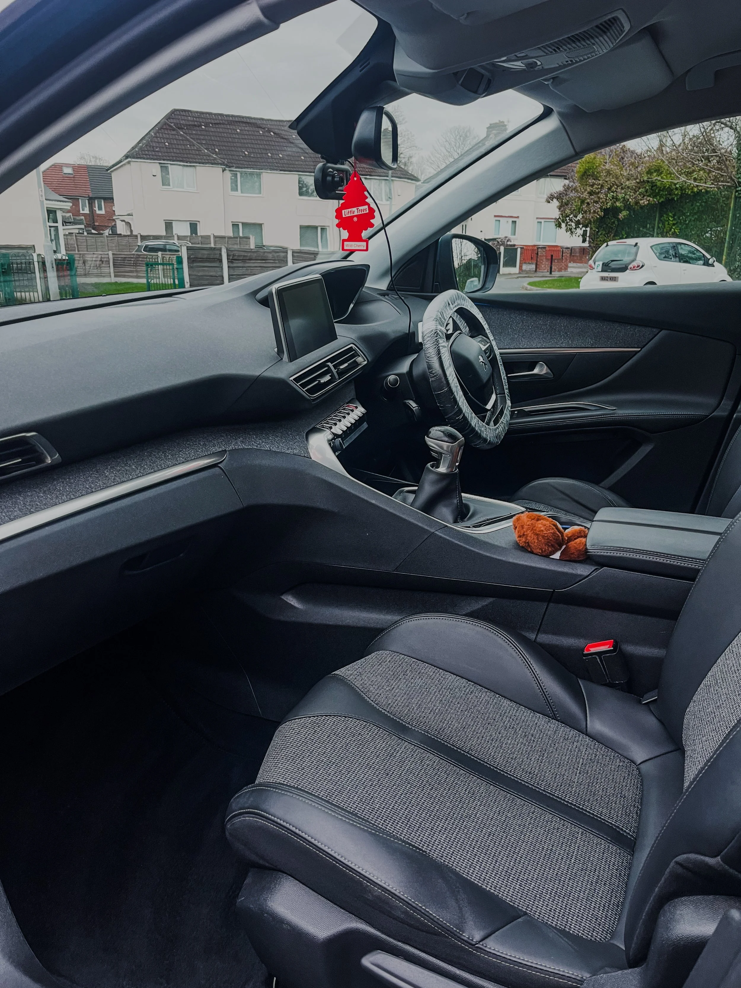 Interior view of a car with a steering wheel, gear shift, dashboard, and front seats, with houses and trees outside the window.