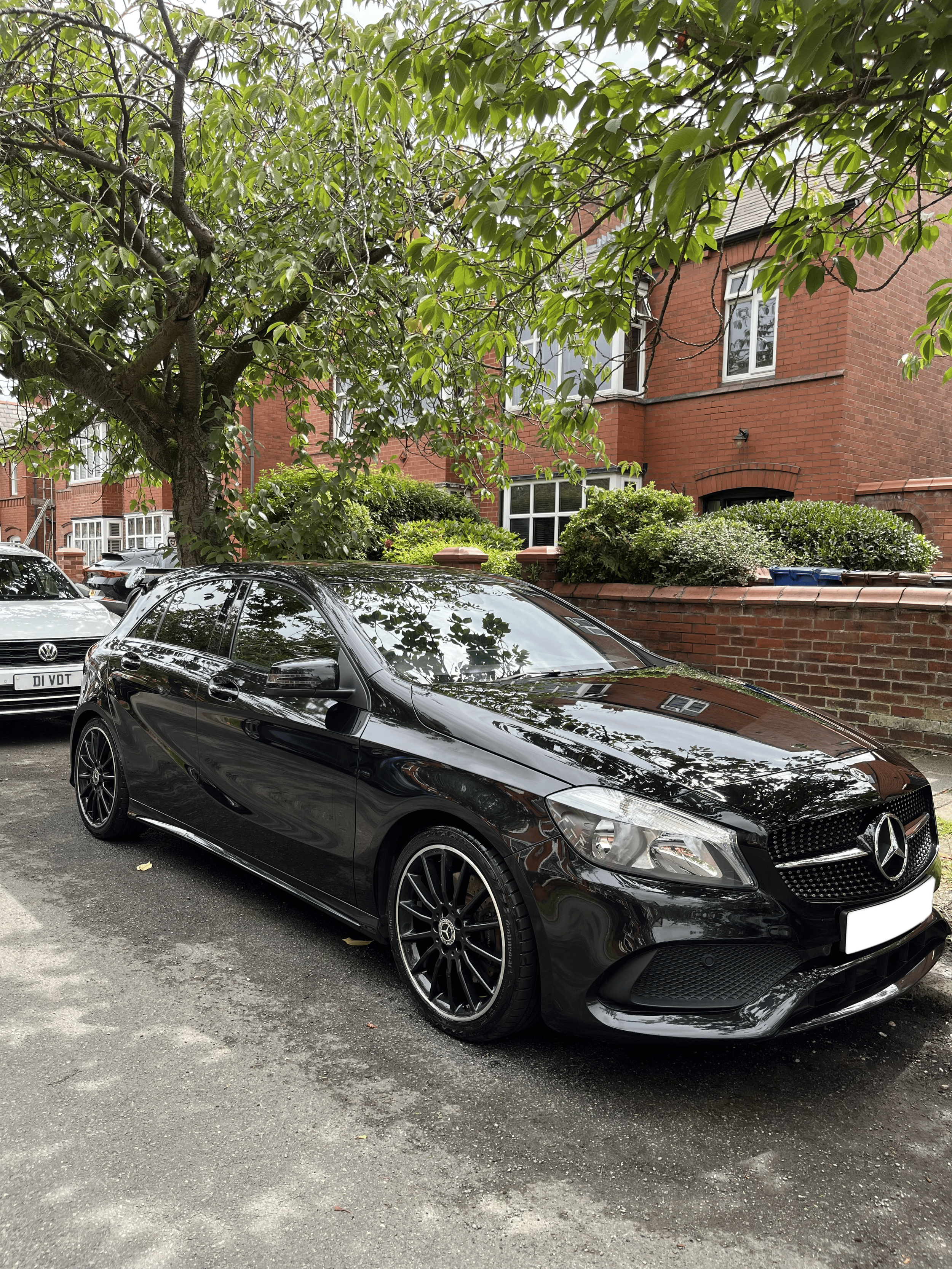 A black Mercedes-Benz hatchback car parked on a residential street, with a tree overhead and red brick houses in the background.
