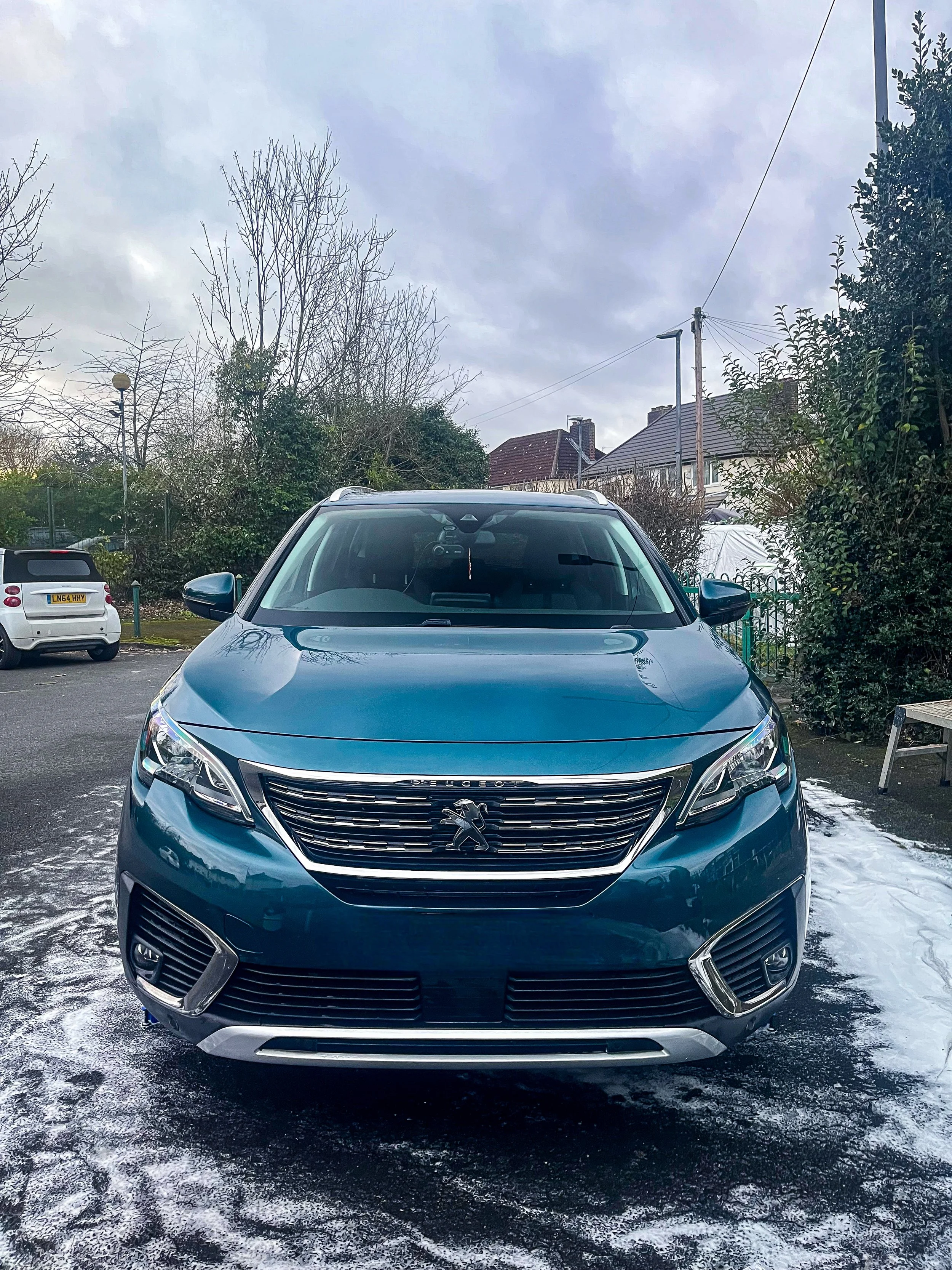 Front view of a blue Peugeot SUV parked outdoors on a snowy surface with trees, houses, and cloudy sky in the background.