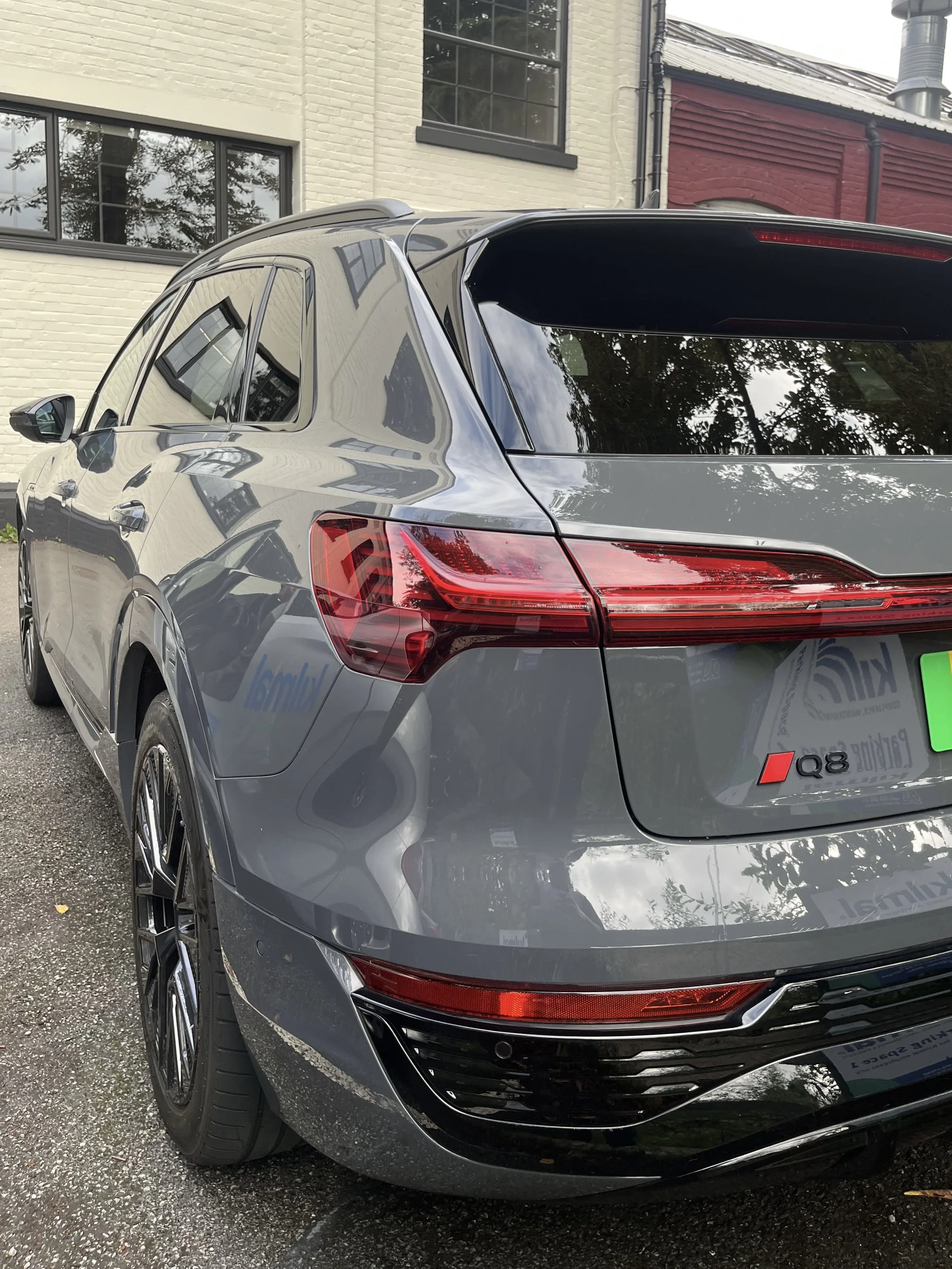 Rear and side view of a gray Audi Q8 SUV parked outdoors on a gravel surface, reflecting nearby buildings and trees, with a residential building in the background.