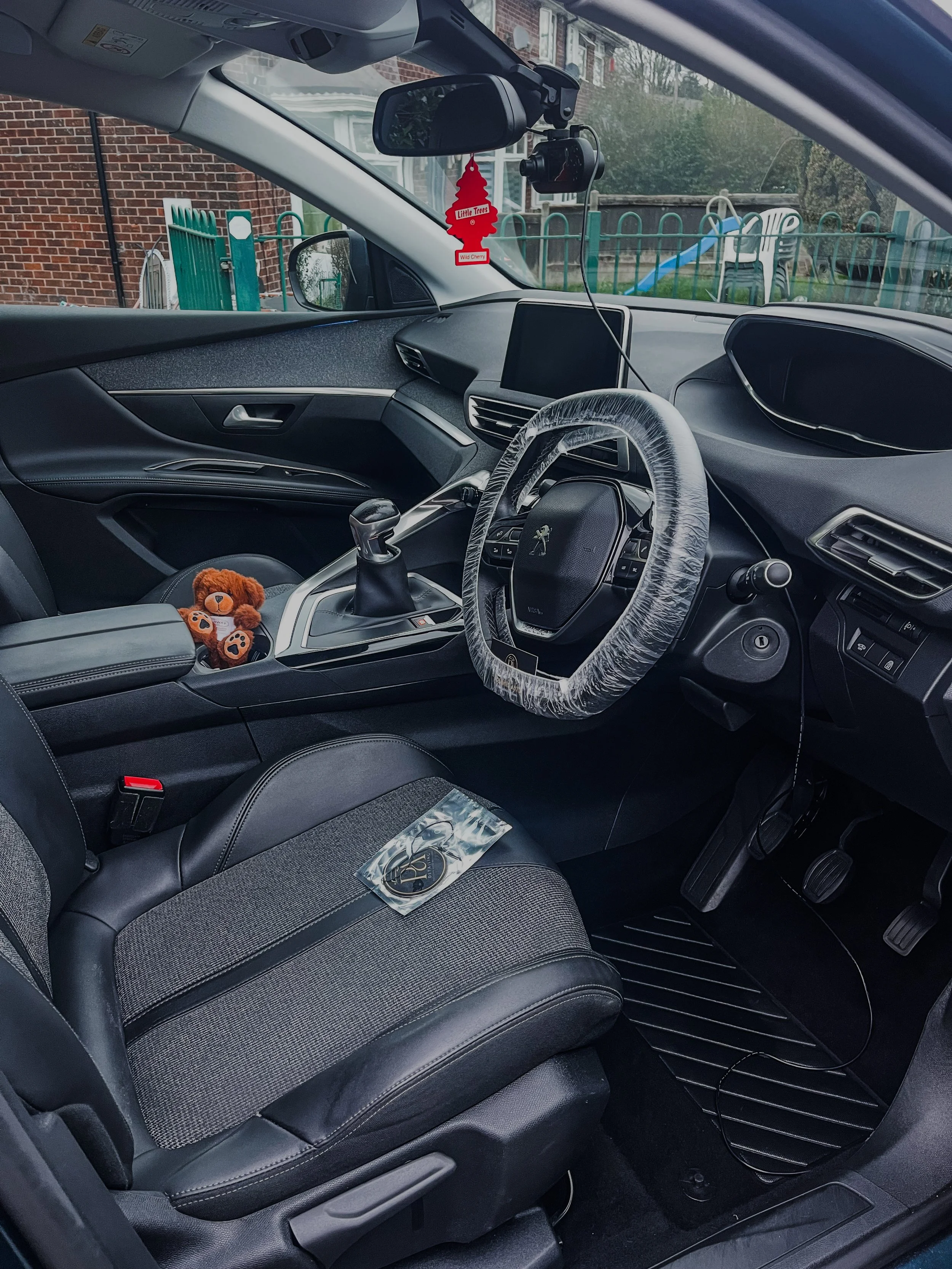 Interior of a car with a stuffed bear on the driver's seat, a gear shift, a steering wheel with a protective cover, a dashboard, a tablet mount, and an air freshener hanging from rearview mirror.