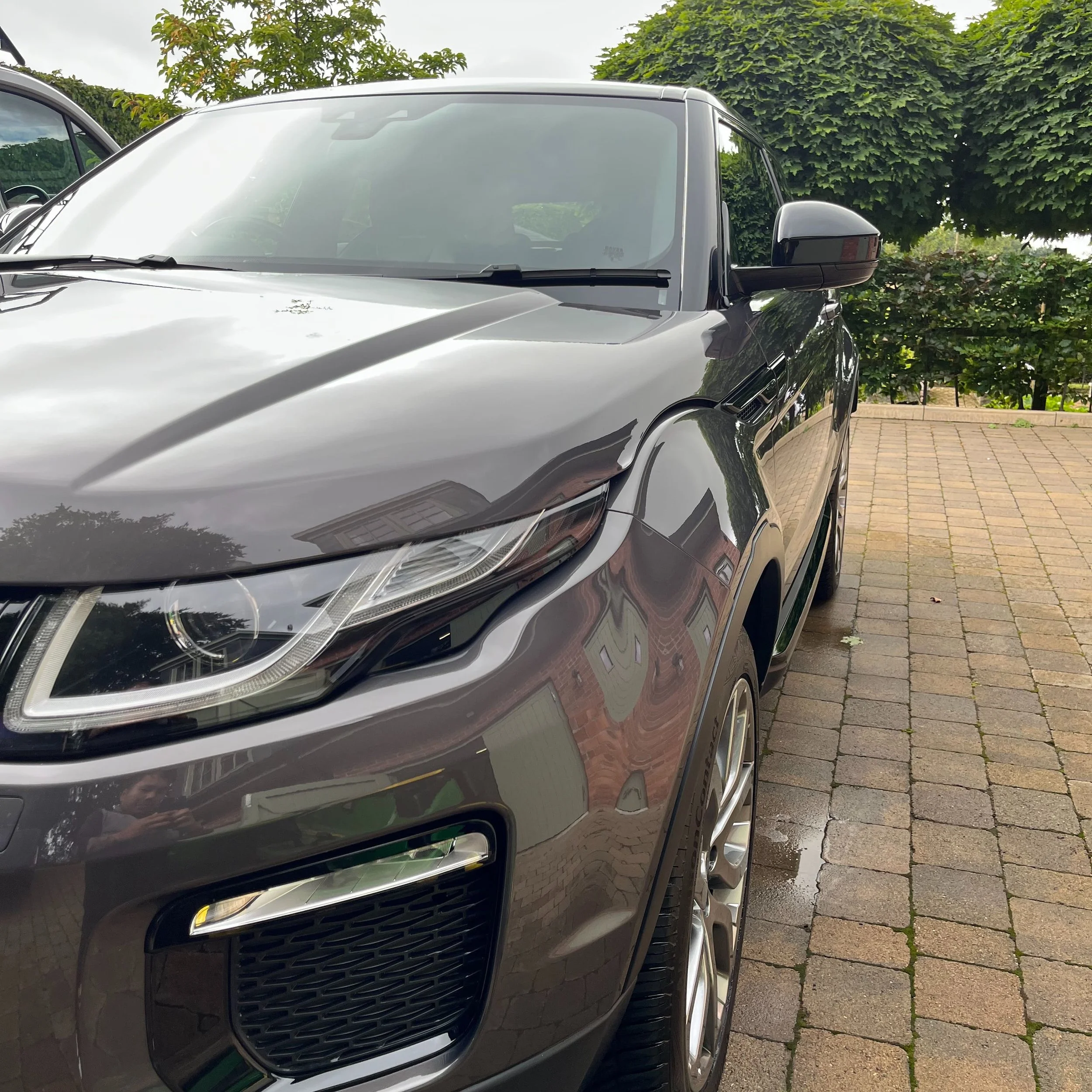 Close-up of a black SUV parked on a brick driveway with trees and bushes in the background.