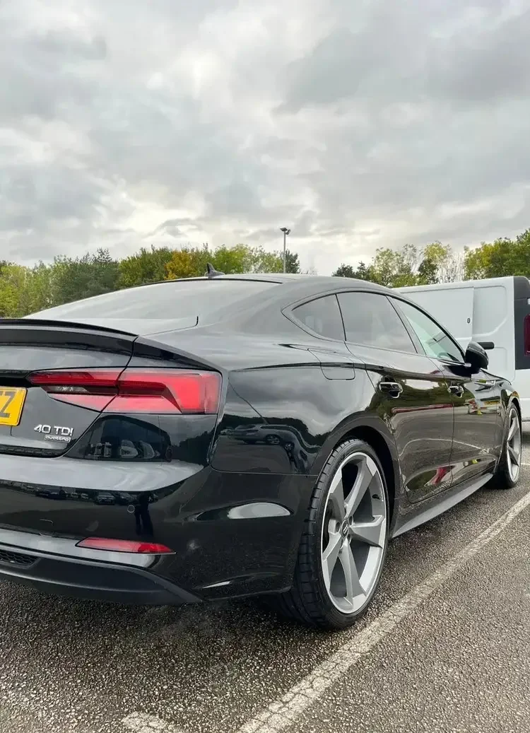 Black Audi A5 Coupe parked in a parking lot with a white van next to it and trees in the background under an overcast sky.