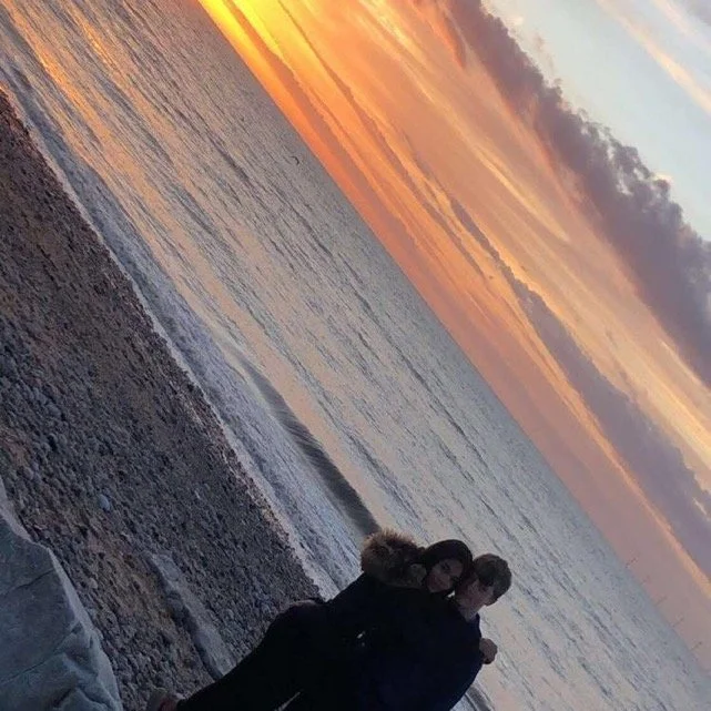 Two people standing on a pebbly beach during sunset, with the sky painted in orange and purple hues over the ocean.