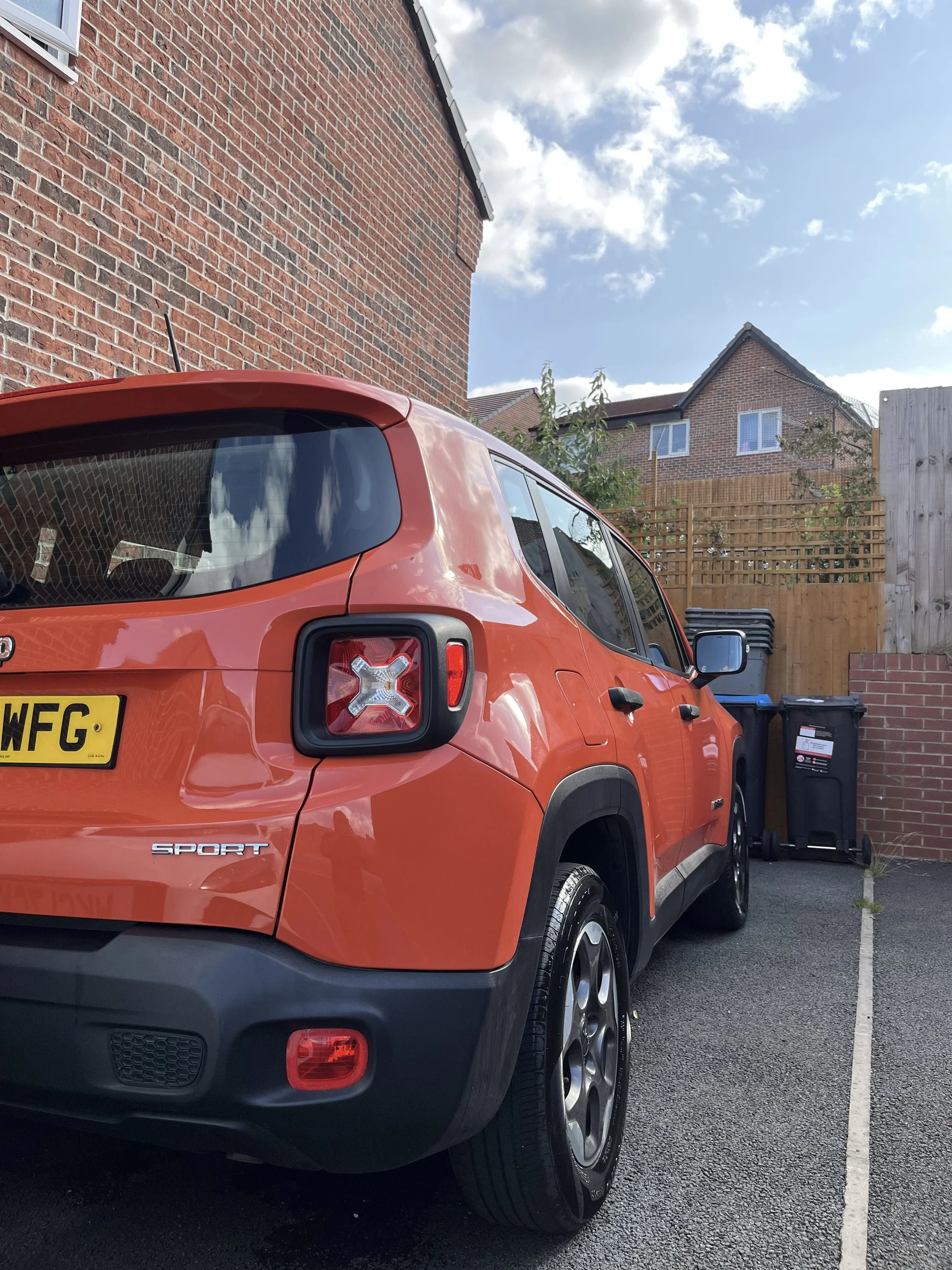 A red Jeep Renegade Sport parked in a driveway next to a brick house, with a wooden fence and trash bins in the background, under a partly cloudy sky.
