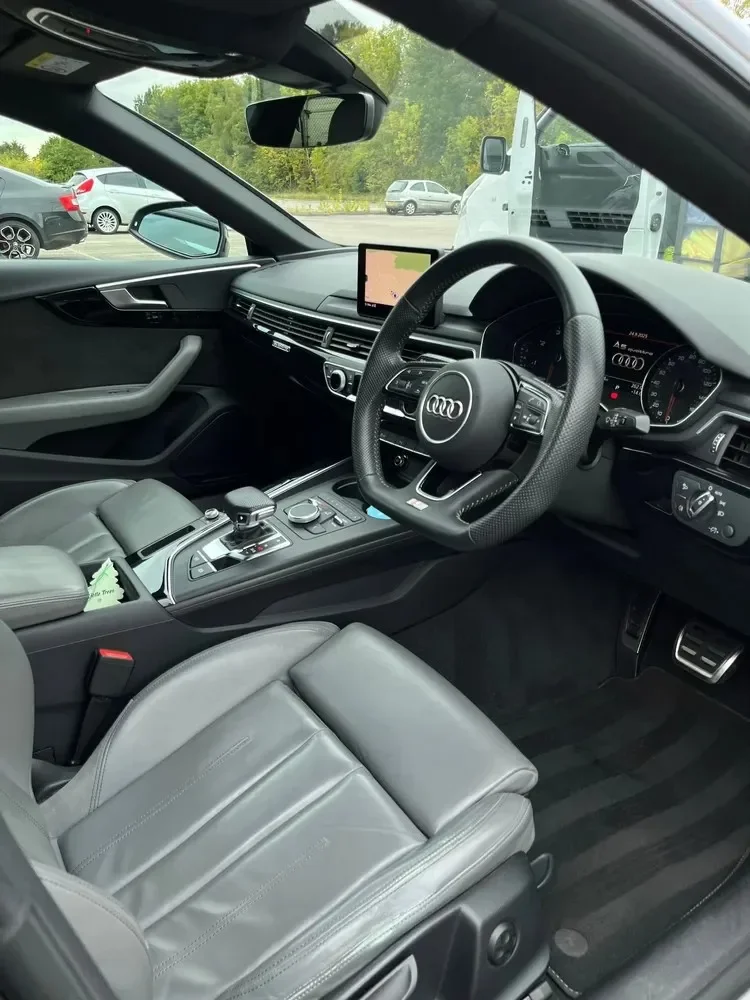 Interior of a modern Audi car showing the steering wheel, dashboard, center console, and front seats. Outside the windshield, there are parked cars and greenery.