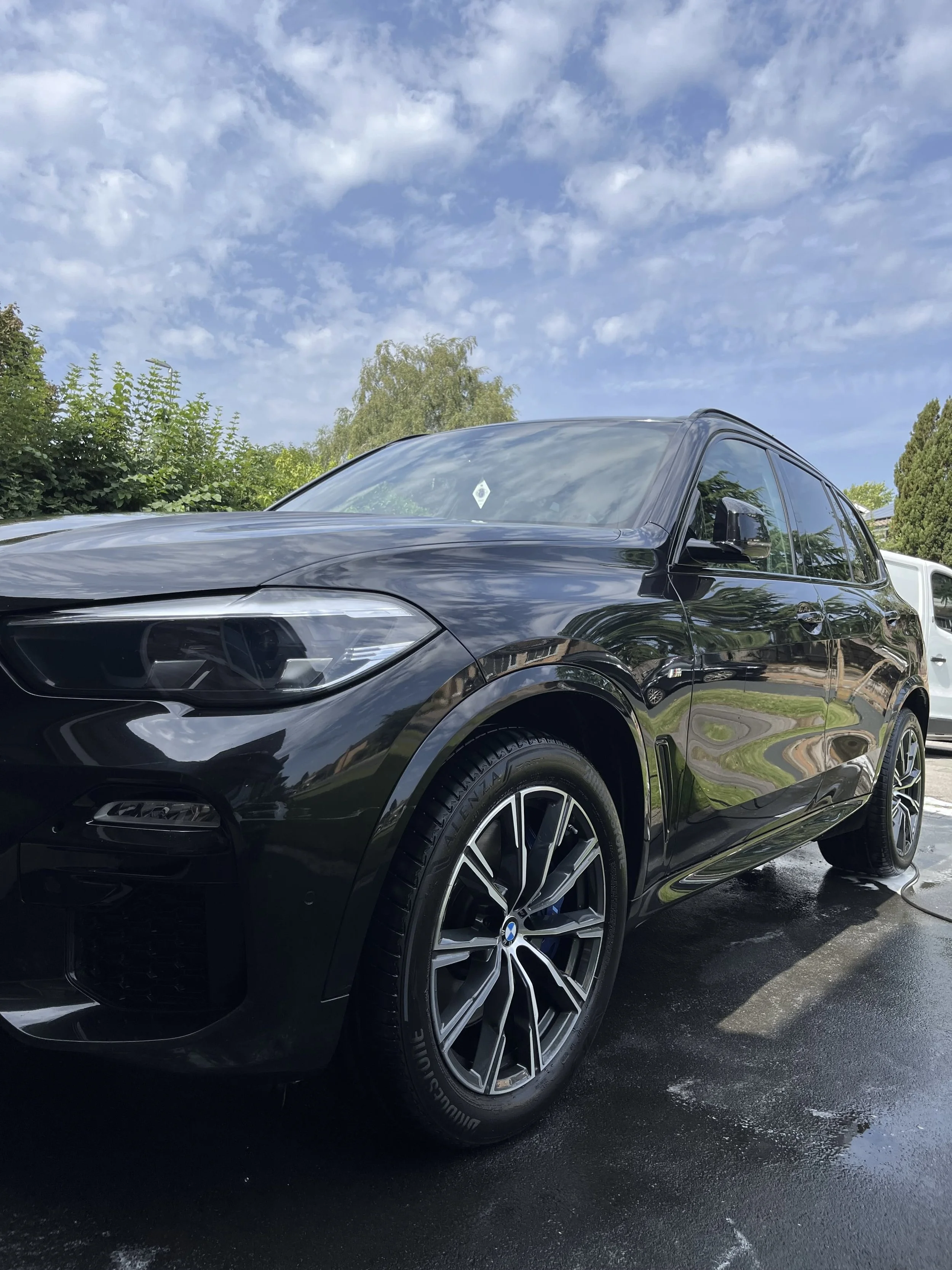 A black BMW parked on a wet surface during daytime, with trees and a partly cloudy sky in the background.