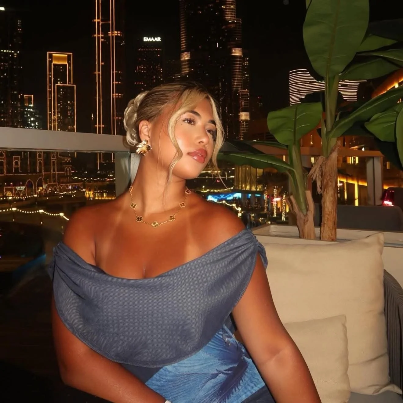 A woman with blonde hair styled in an updo and loose curls framing her face, wearing a off-shoulder blue dress, gold jewelry including earrings and necklace, sitting on a lounge chair against a city skyline at night with illuminated skyscrapers and bright city lights.