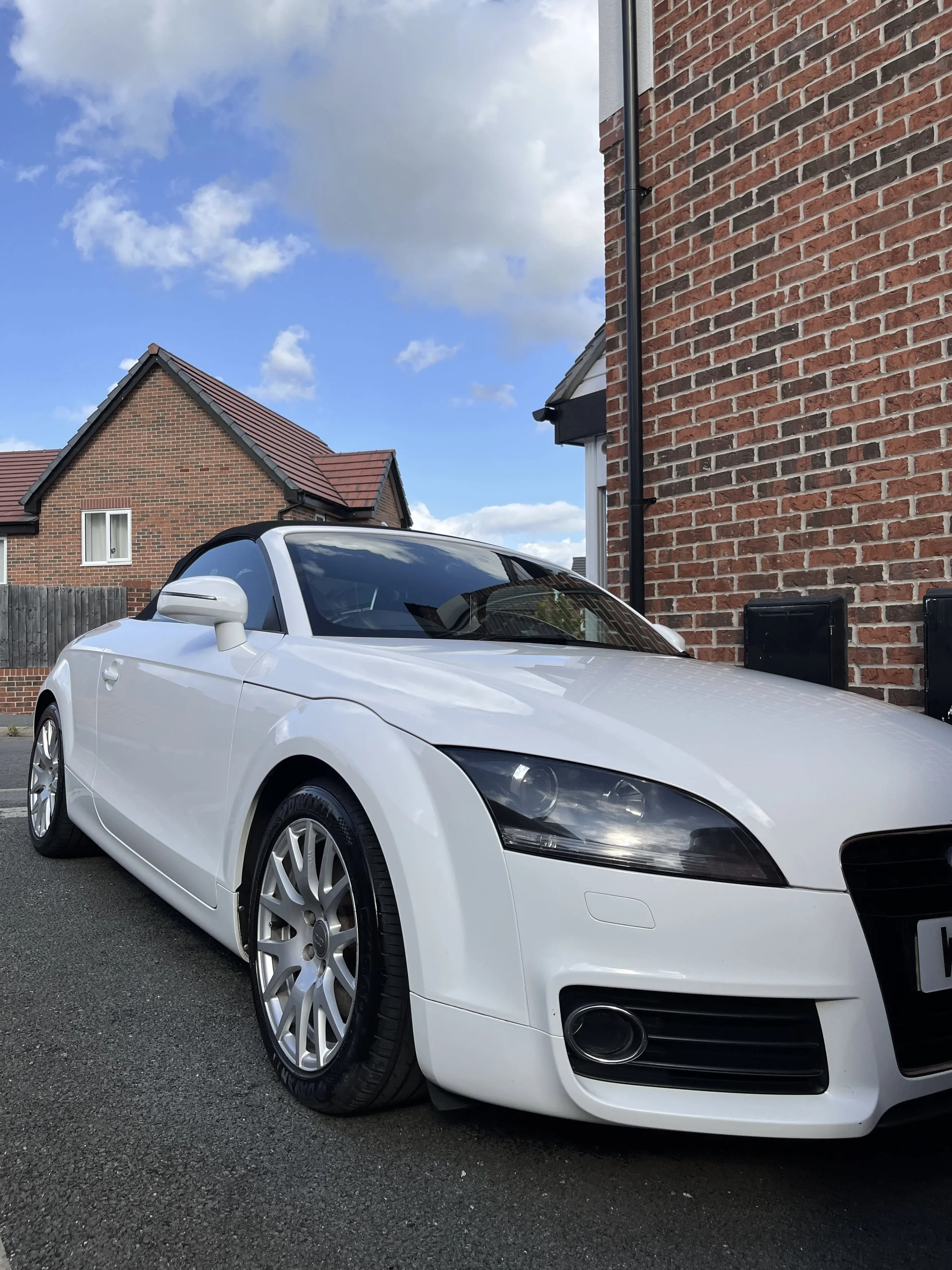White convertible car parked next to a brick house under a partly cloudy sky.