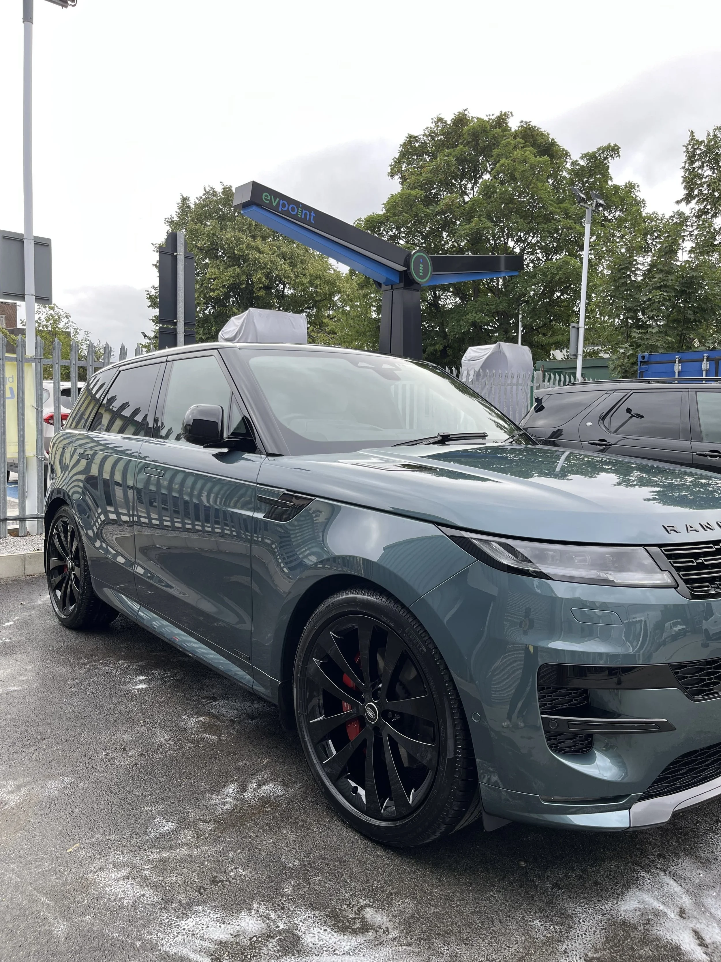 A metallic gray Range Rover parked at an electric vehicle charging station with a blue and black sign reading 'ev point' in front of trees and cloudy sky.