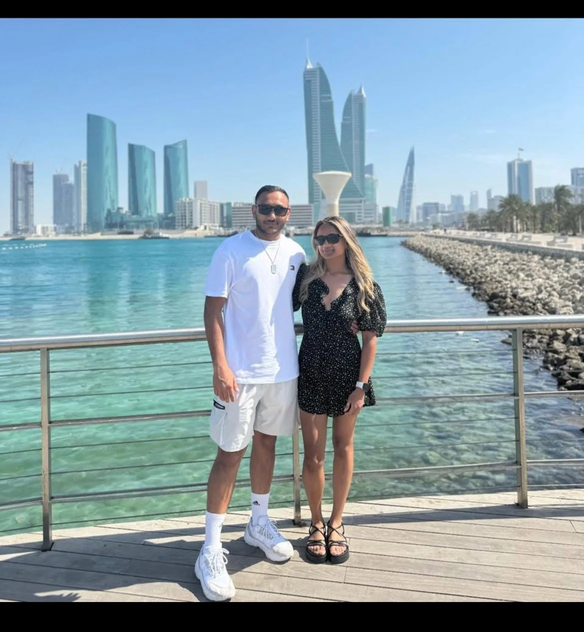 A man and woman standing side by side on a pier overlooking a harbor with modern skyscrapers in the background, sunny sky, and clear water.