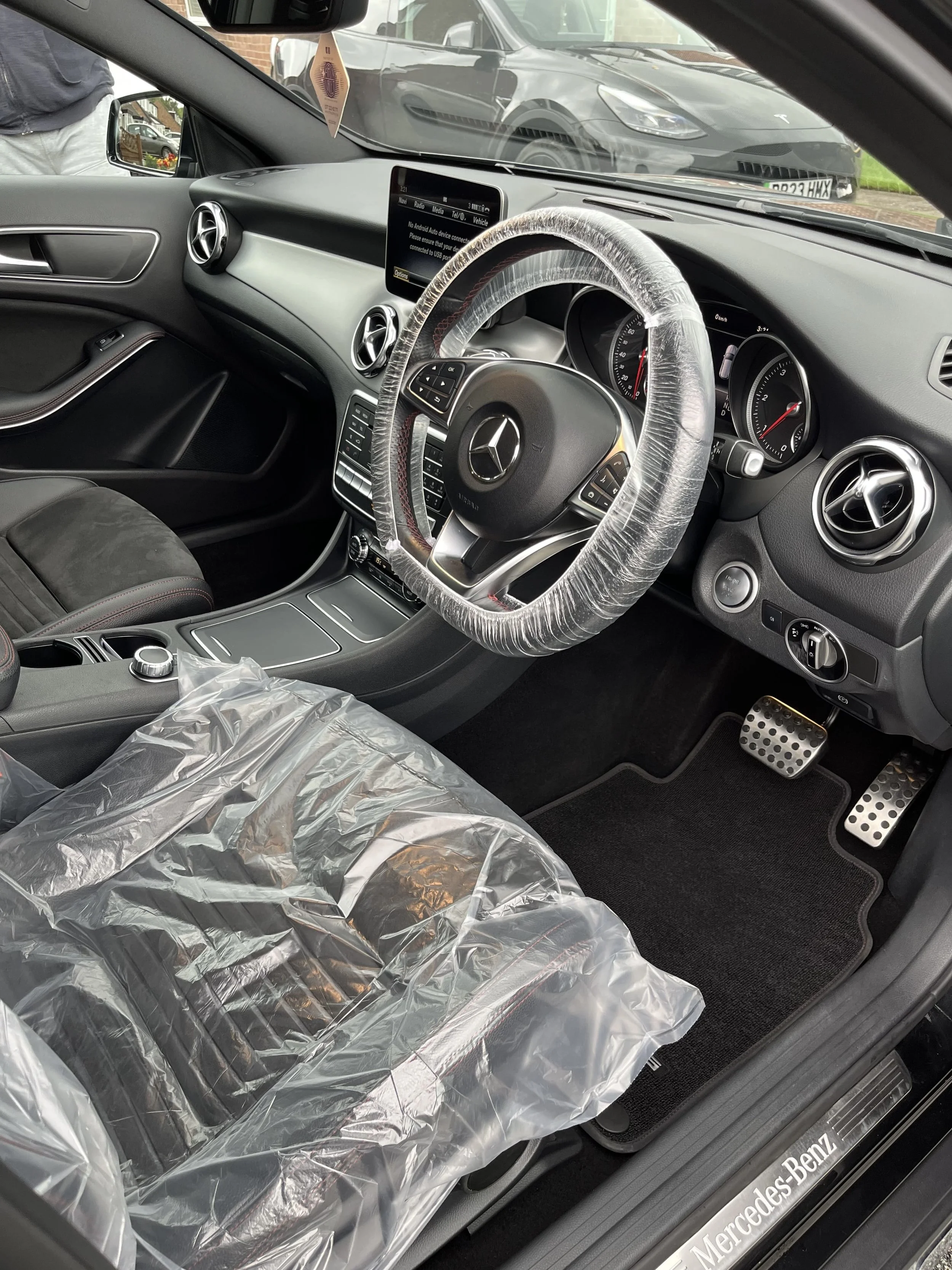 The interior of a new Mercedes-Benz car, with a plastic cover on the driver's seat and a wrapped steering wheel, parked outside a parking lot with other cars visible through the windshield.