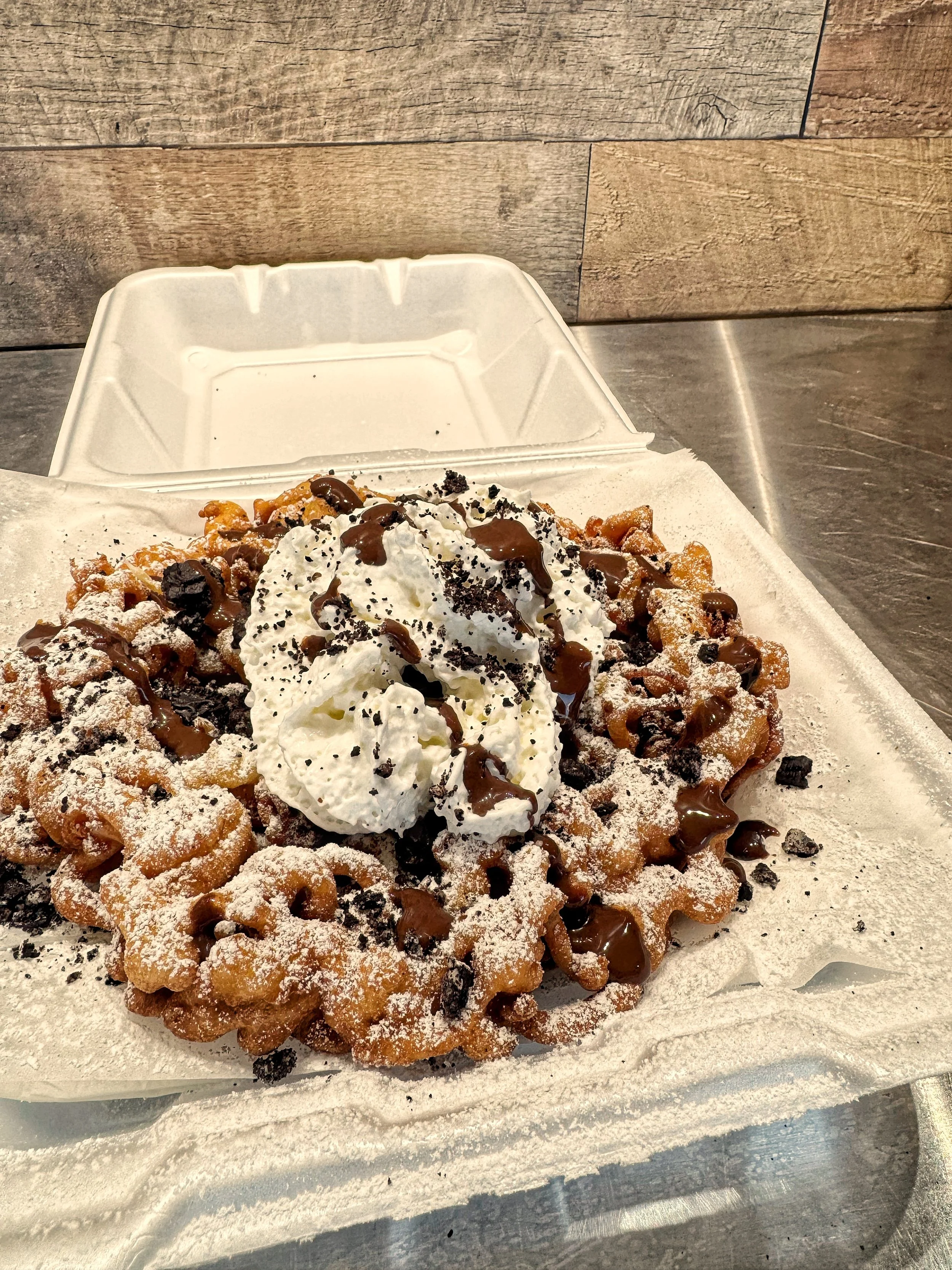 Close-up of a Funnel Cake topped with whipped cream, chocolate syrup, and cookie crumbs on parchment paper.