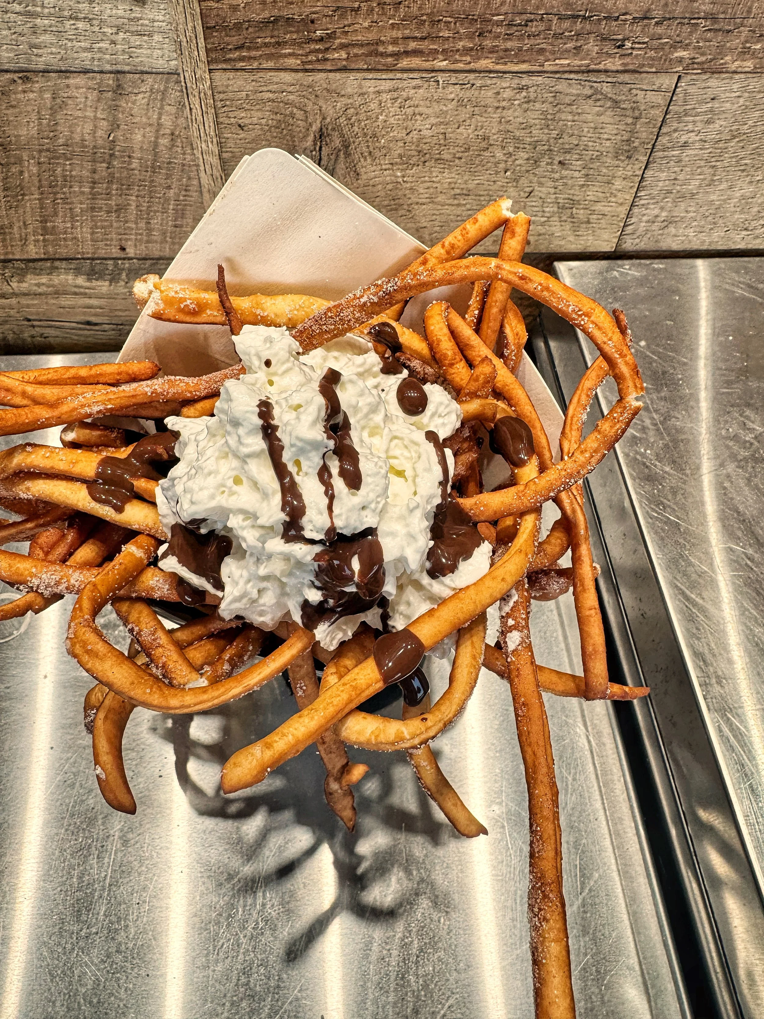 A serving of loaded Elephant fries topped with whipped cream, and chocolate syrup on a metal tray.