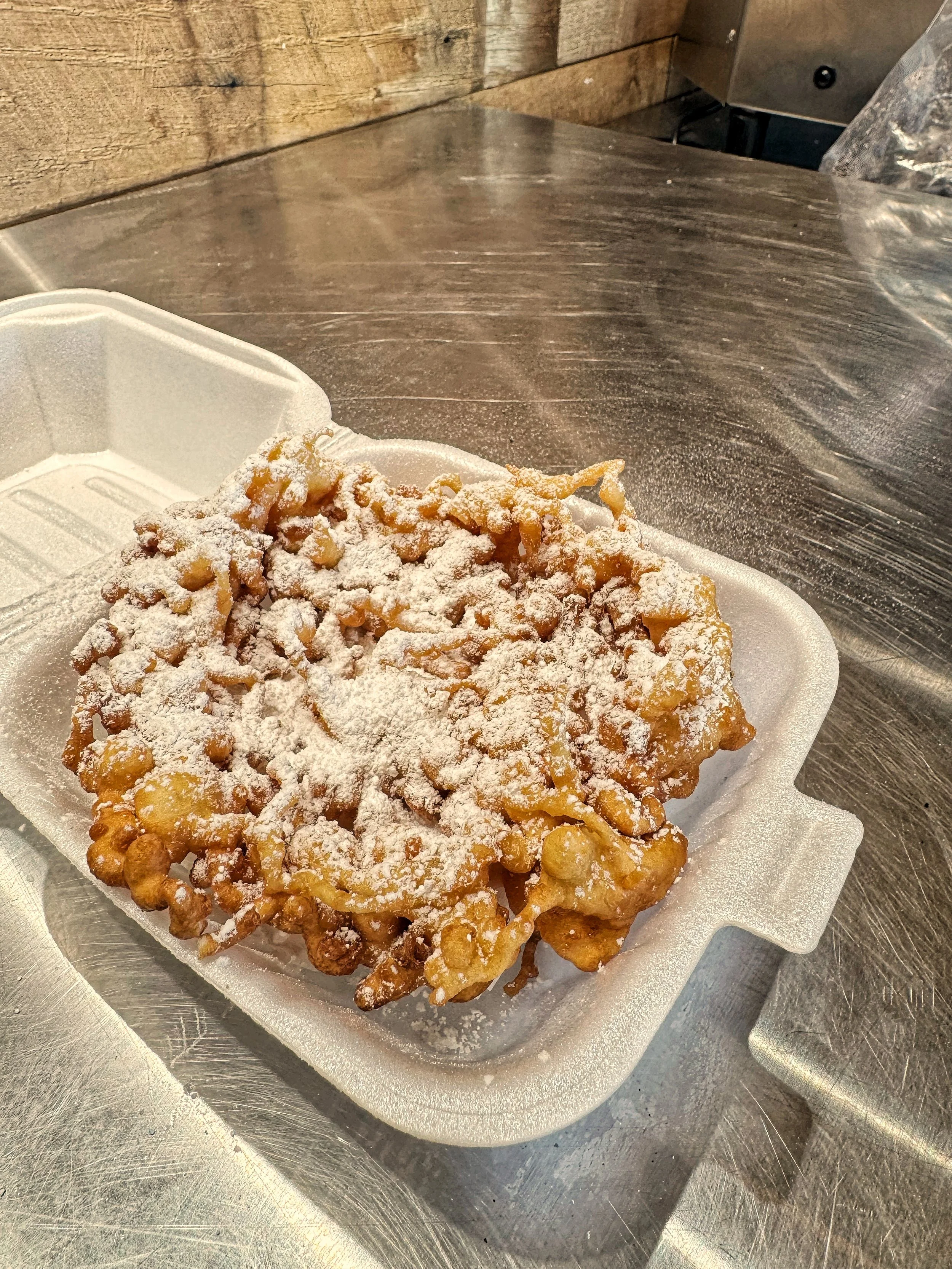 A funnel cake topped with powdered sugar on a foam tray, on a stainless steel counter.