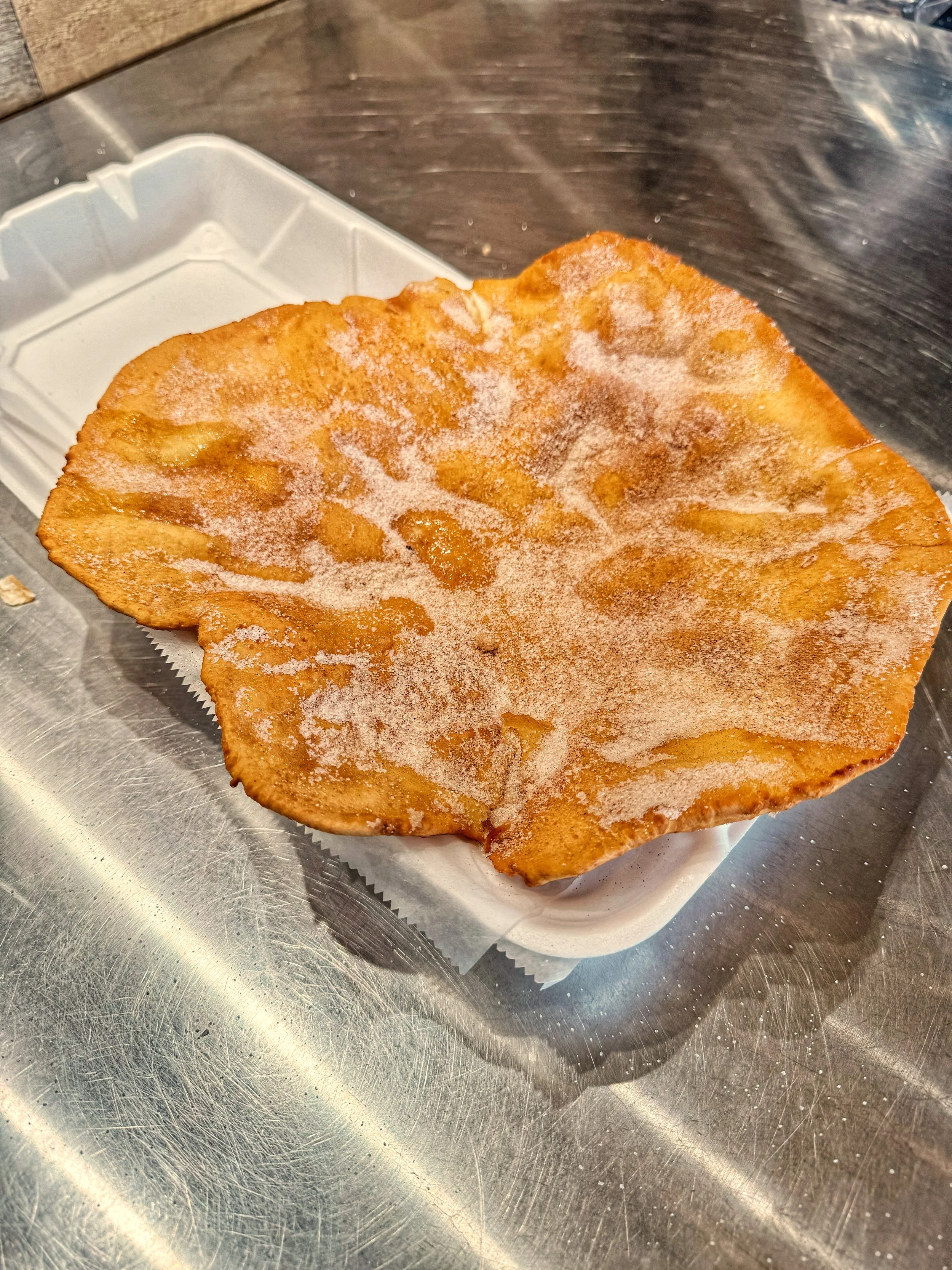 A elephant ear topped with sugar, and cinnamon, placed on a paper-lined tray on a stainless steel countertop.