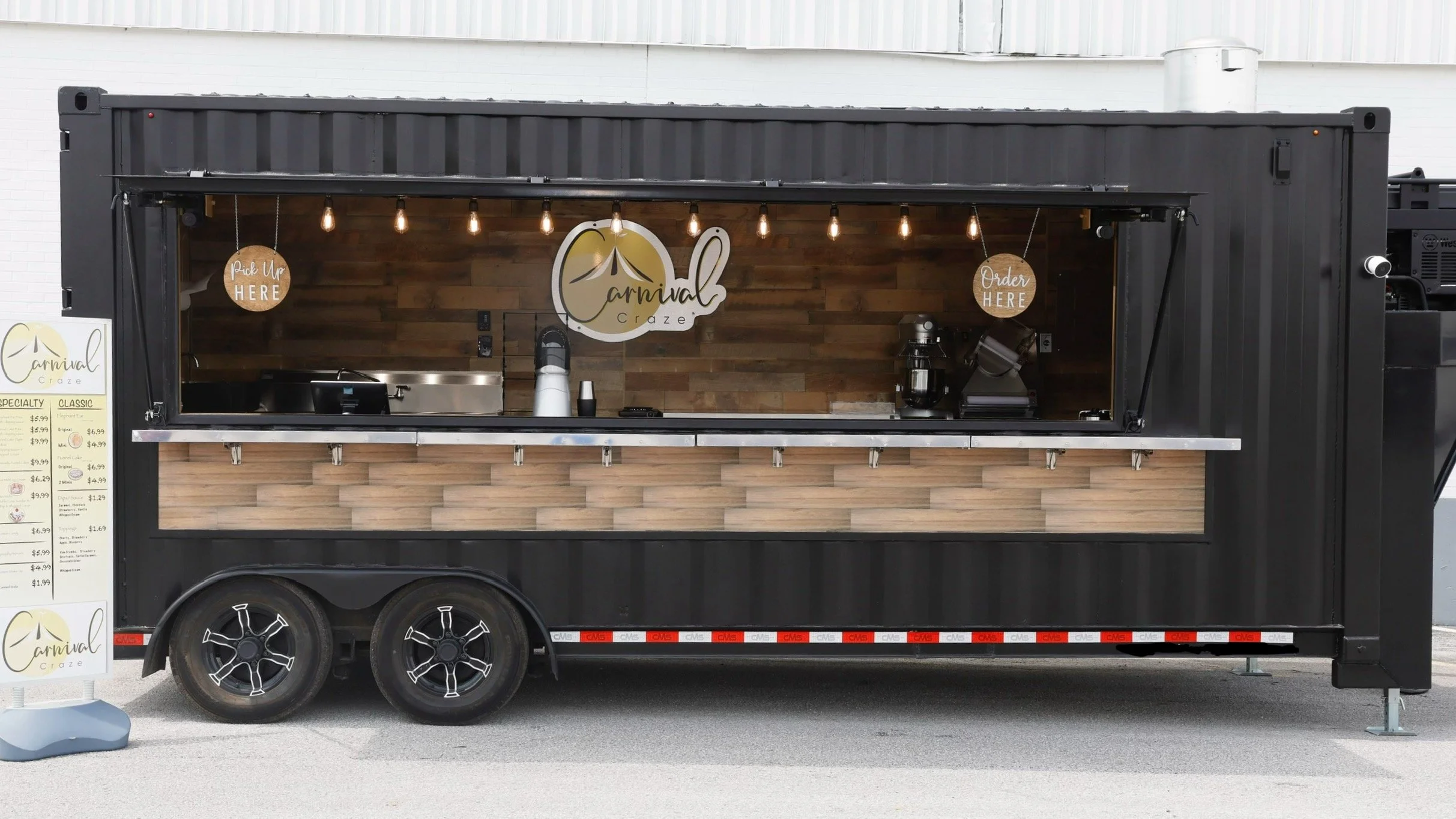 A black mobile food truck with a wooden counter and a sign that reads 'Carnival Craze' serving food and beverages, with lights hanging above.