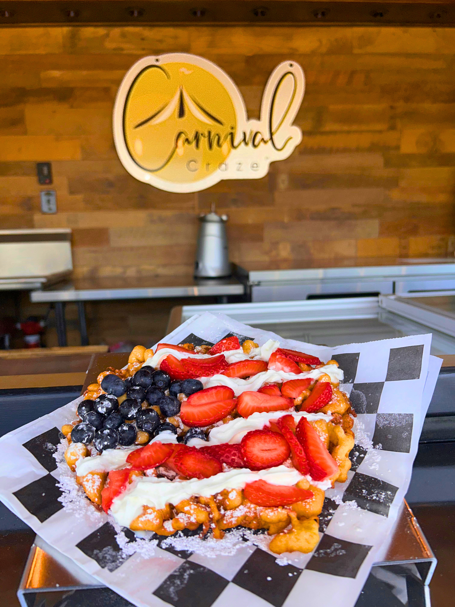 A dessert waffle topped with strawberries, blueberries, whipped cream, and caramel syrup, sitting on black and white checkered paper lined tray.