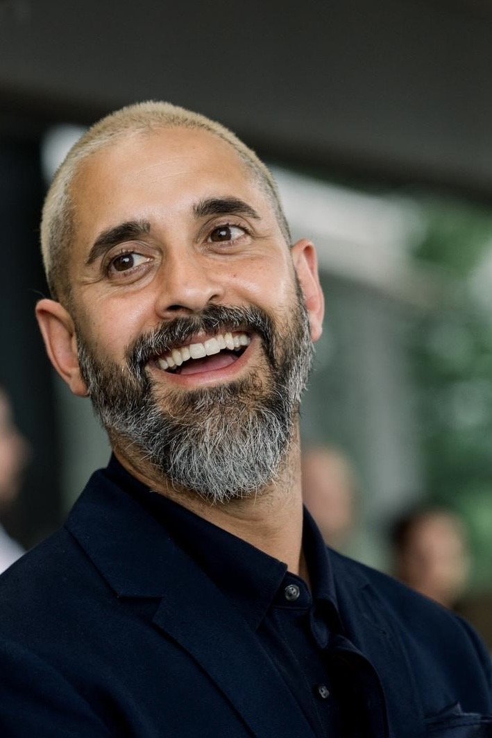 A smiling man with a beard and short, bleached blonde hair, wearing a dark suit and shirt, in an indoor setting with blurred background.