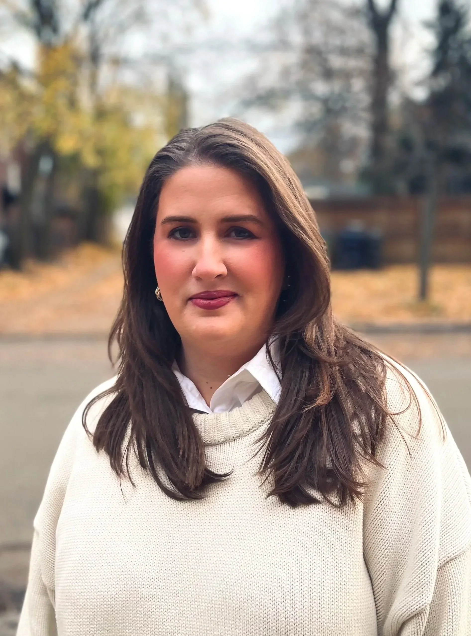 A woman with long dark hair wearing a cream sweater over a white collared shirt, standing outdoors with blurred trees and a paved area in the background.