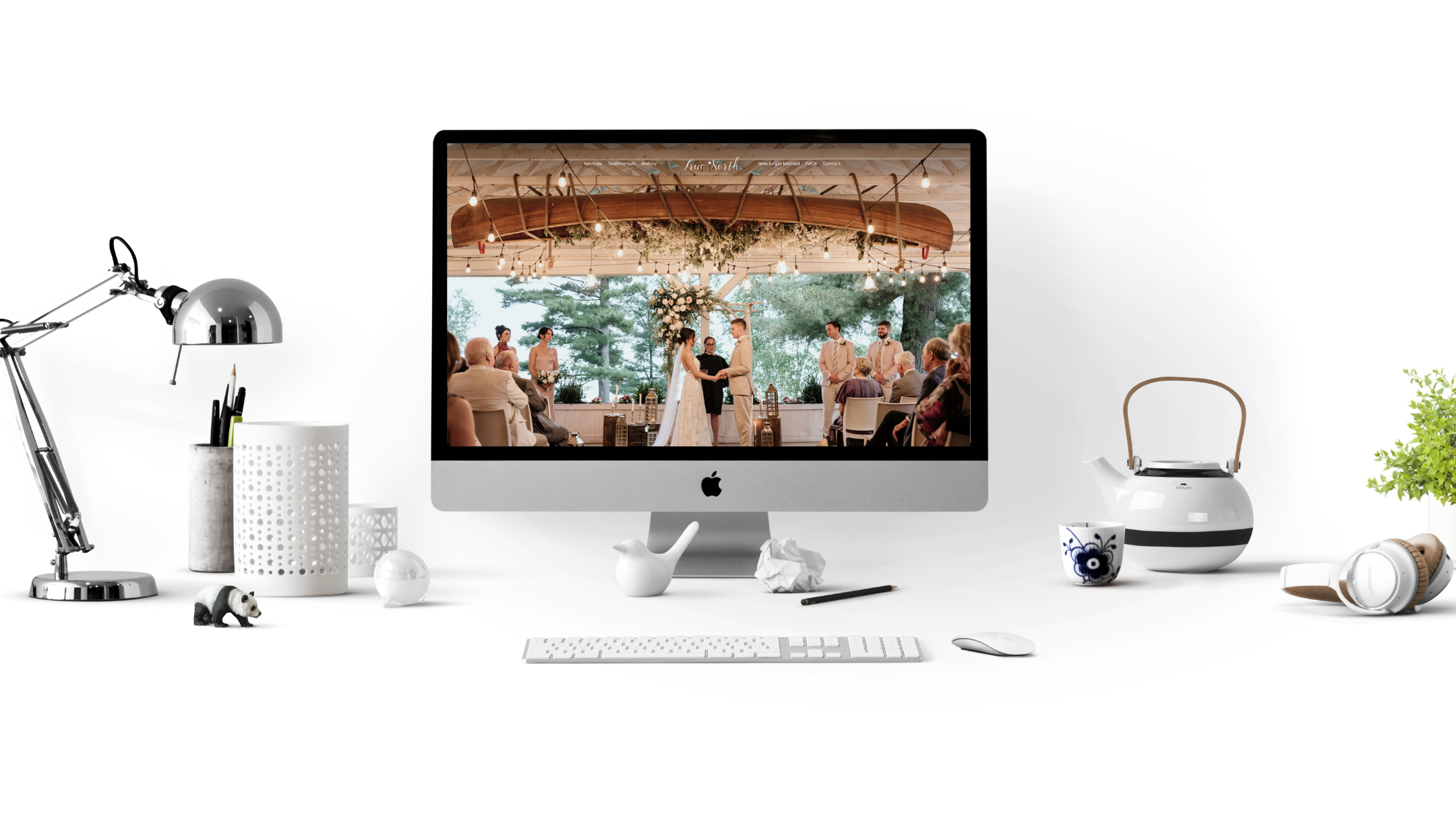 A desktop computer with a wedding ceremony displayed on the screen. The desk also features a lamp, pens in a holder, a teapot, a cup, headphones, and a small plant.
