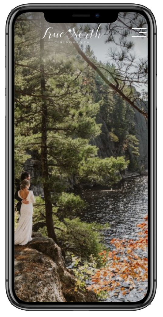 Couple in wedding attire standing on a rock ledge surrounded by trees near a lake, viewed on a smartphone screen.