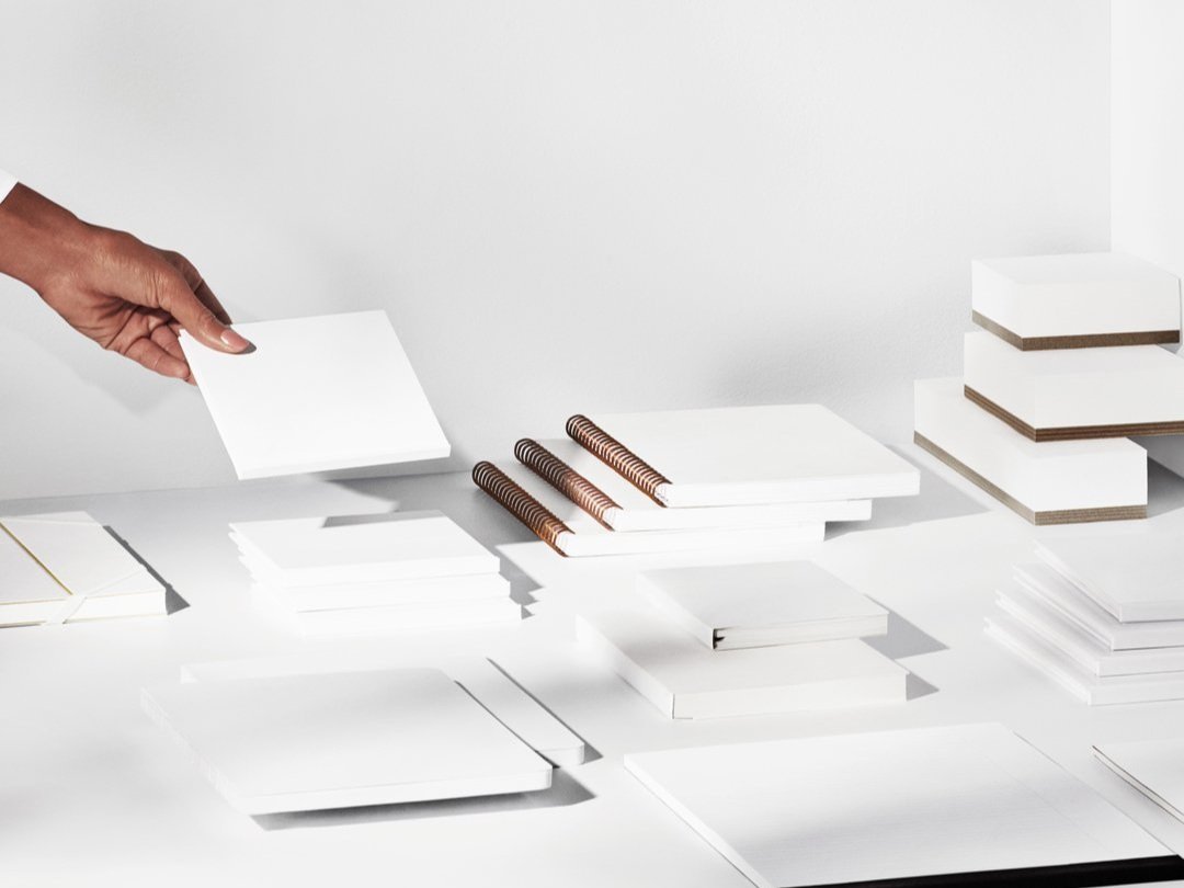 Person arranging white stationery items on a table, including stacks of notepads, spiral notebooks, and envelopes, with a minimalist design.