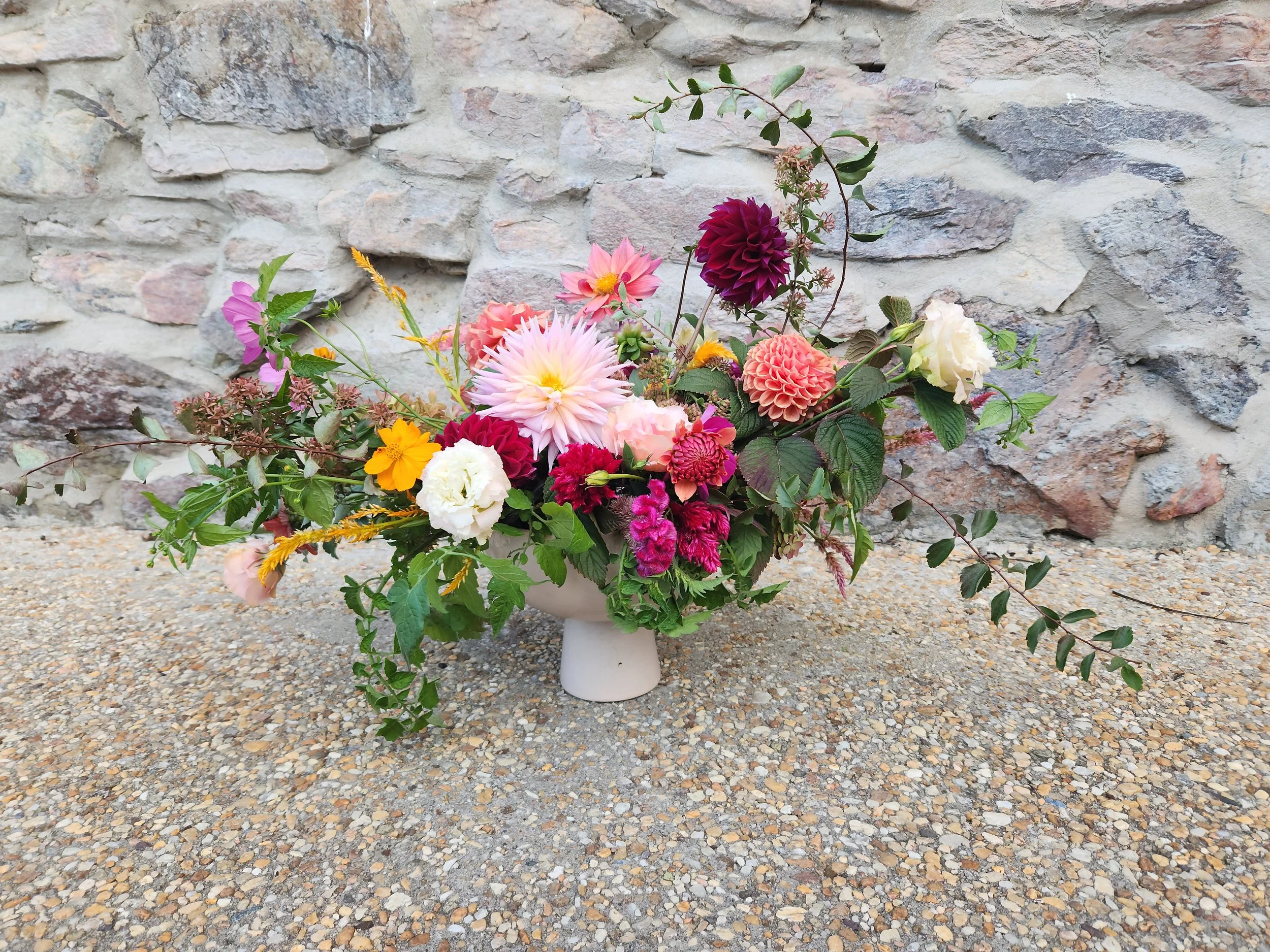 Colorful flower arrangement in a white vase placed on a gravel surface against a stone wall background.