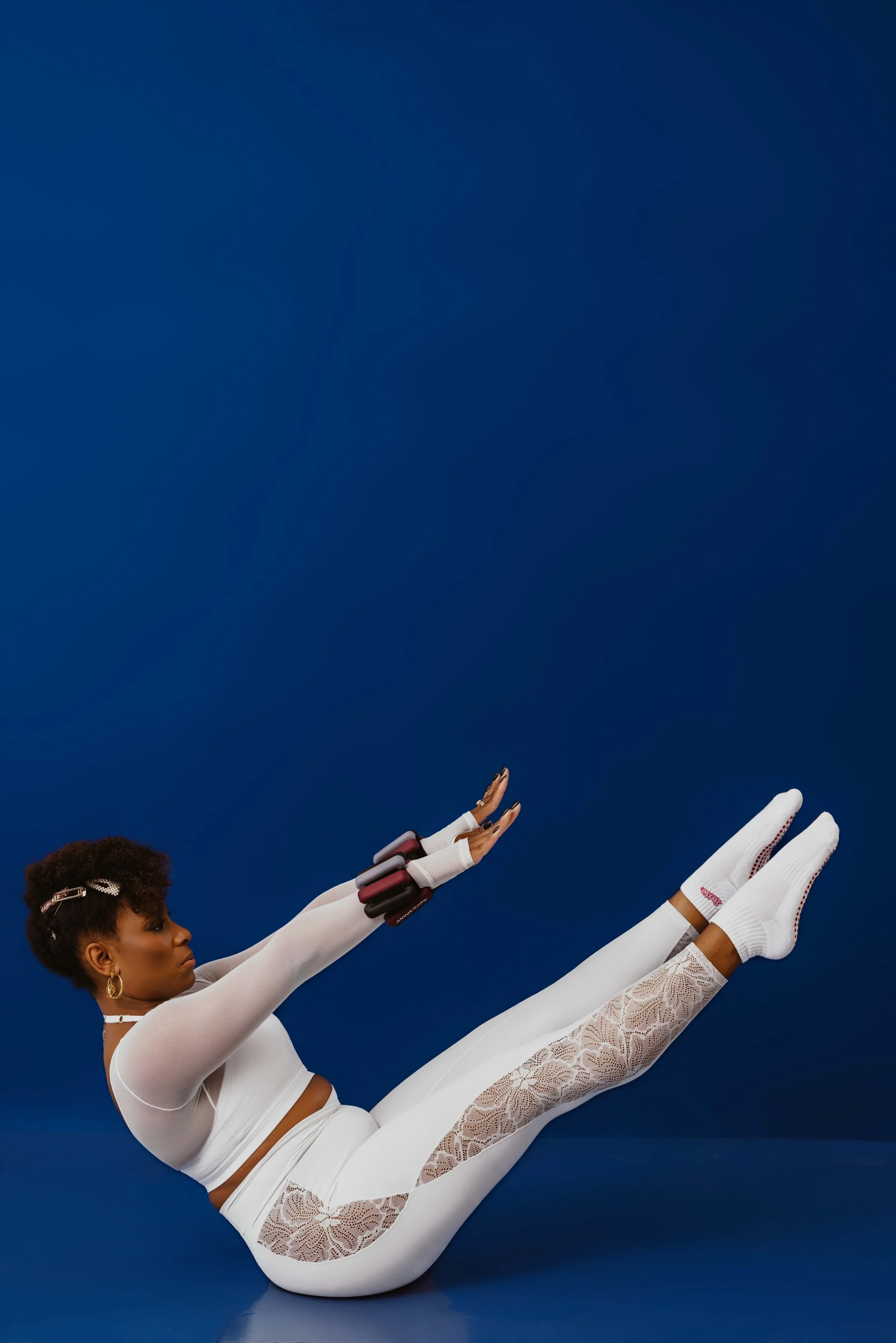 A woman in white athletic clothing, with lace details and white shoes, is lying on her back with her legs elevated and extended against a dark blue background. She has short, curly hair adorned with a bow and is reaching upward with her right hand, which is decorated with a black and red fitness tracker, while wearing white gloves.