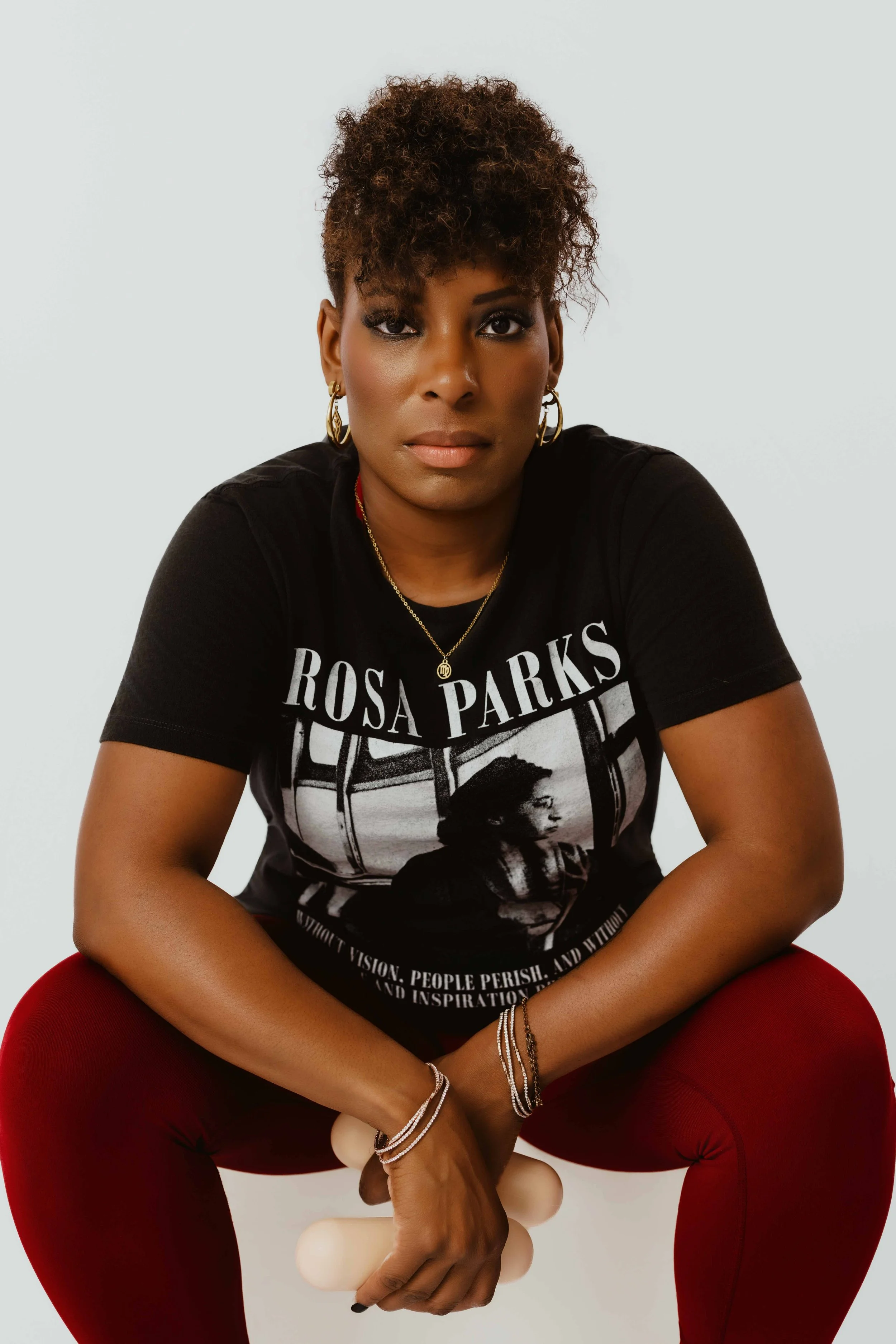 A woman with short curly hair, wearing makeup, gold earrings, a necklace, and a black graphic T-shirt, sitting on a red chair with legs crossed, holding a beige object, against a plain white background.