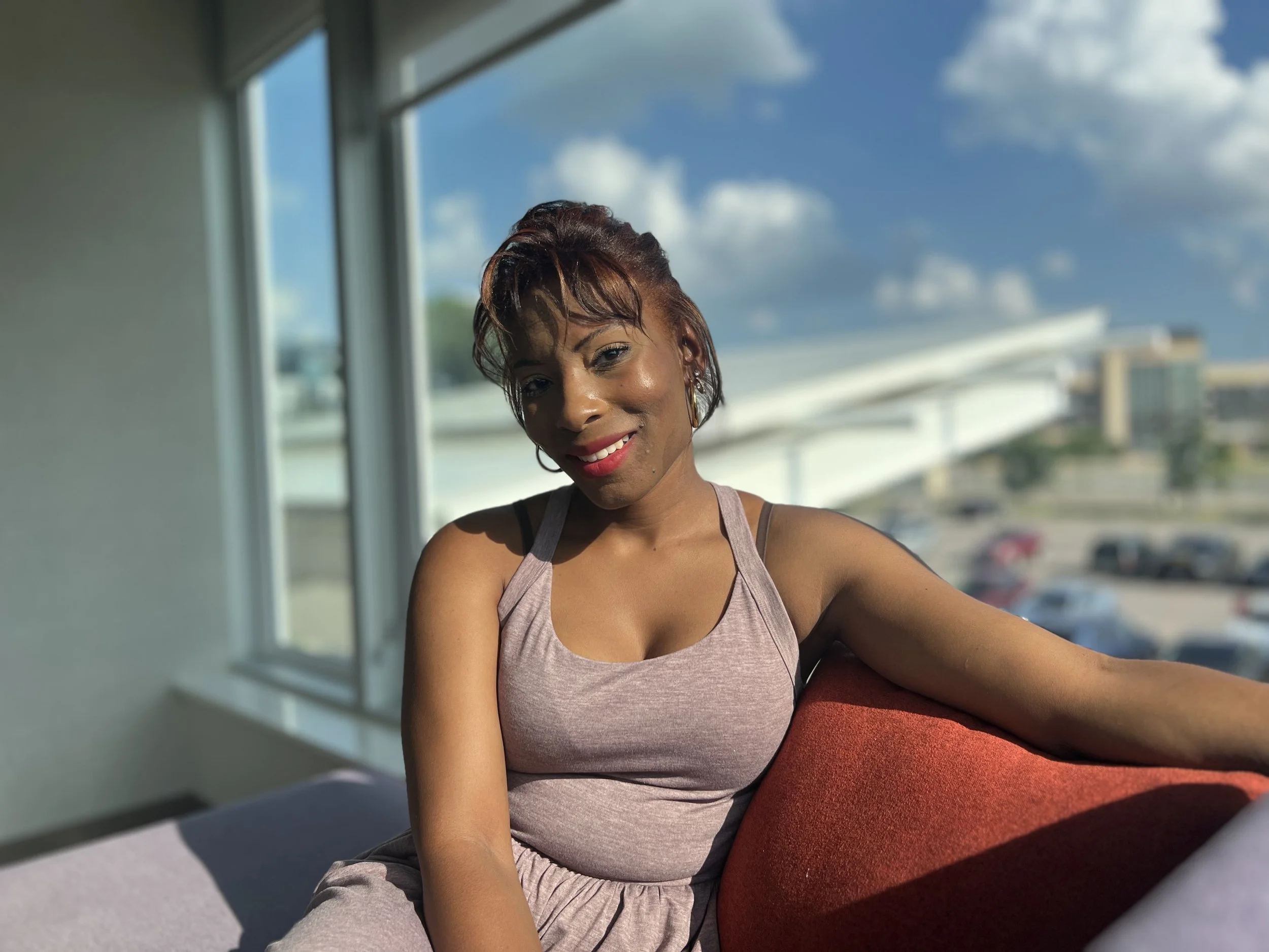 A smiling Black woman with short reddish-brown hair and earrings sitting on a red couch, wearing a light pink tank top, with a large window behind her showing a partly cloudy blue sky and a parking lot.