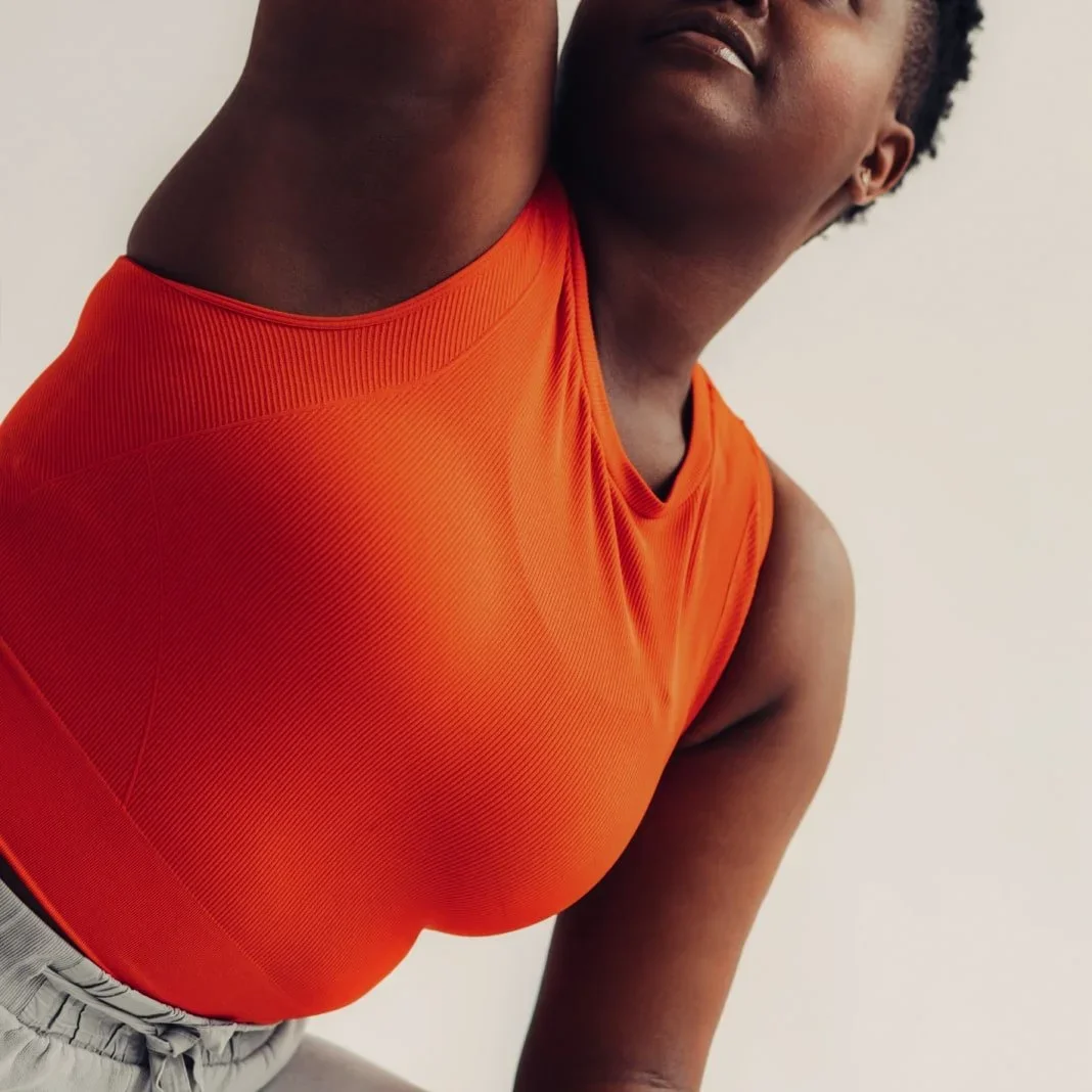Close-up of a Black woman in an orange athletic top, capturing her torso and part of her face as she leans to the side.