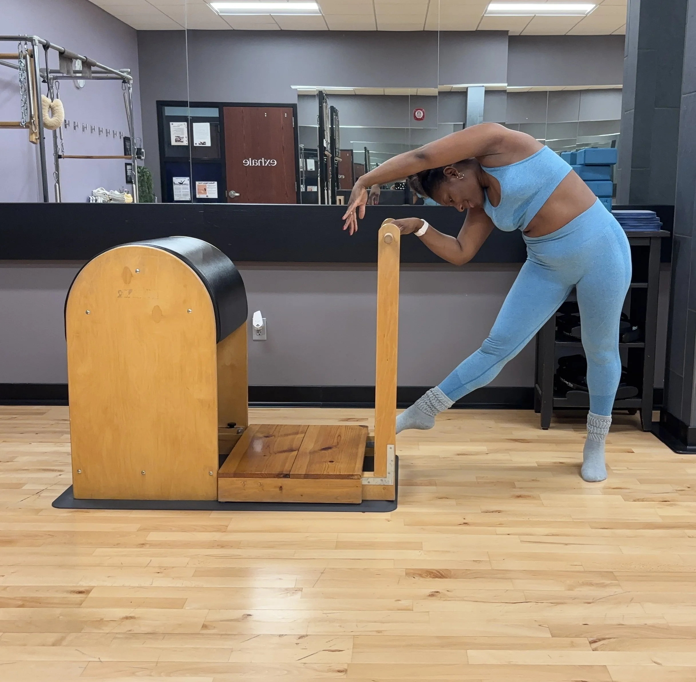 A Black woman in blue workout clothes stretching at a wooden Pilates reformer machine in a gym with wood flooring, mirrors, and fitness equipment.