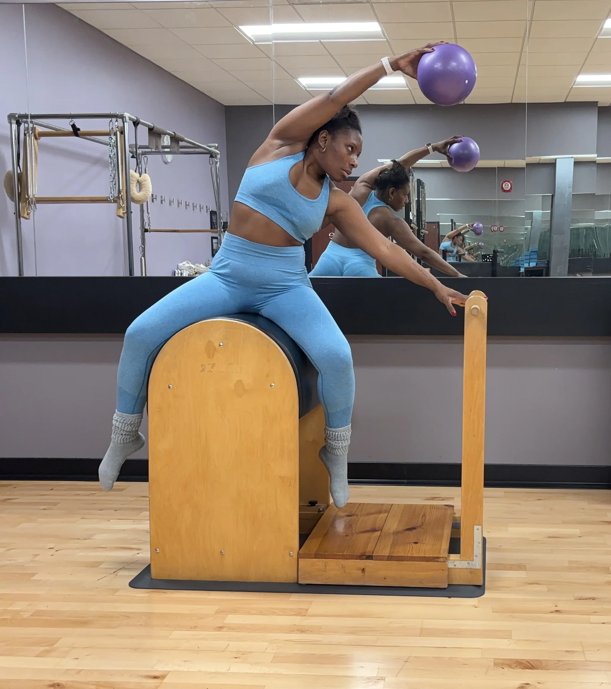 A Black woman in blue workout attire yoga poses on the ladder barrel to promote balance in Pilates in the studio.