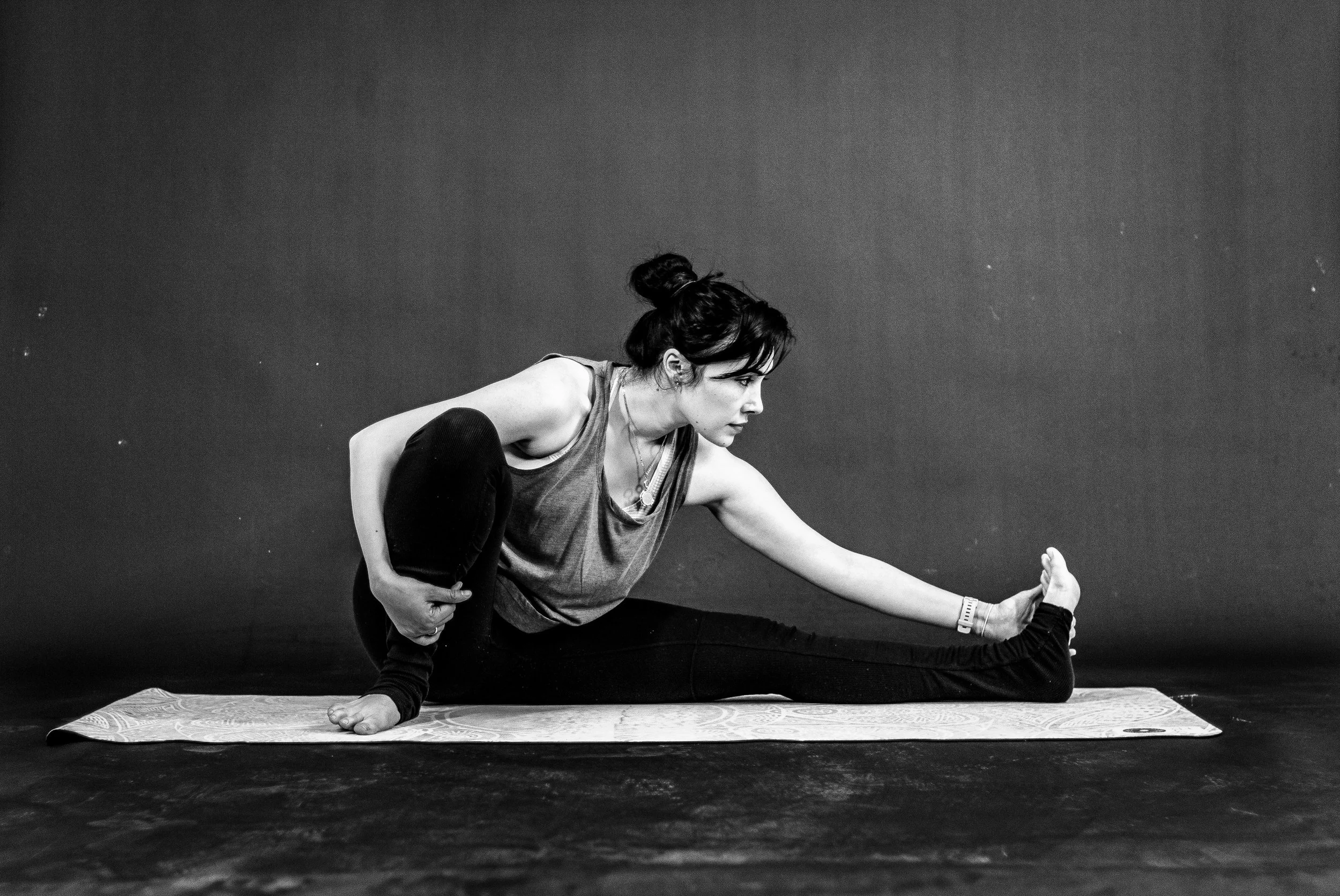 A woman practicing yoga on a mat, sitting with one leg extended and the other folded, reaching toward her foot, in a black and white photo.