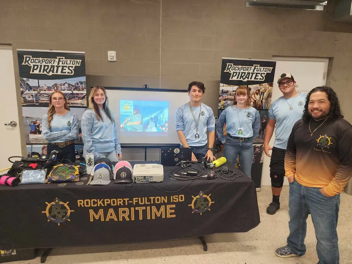 Group of six people standing behind a table with maritime equipment, with banners for Rockport-Fulton Pirates in the background, indoor setting.