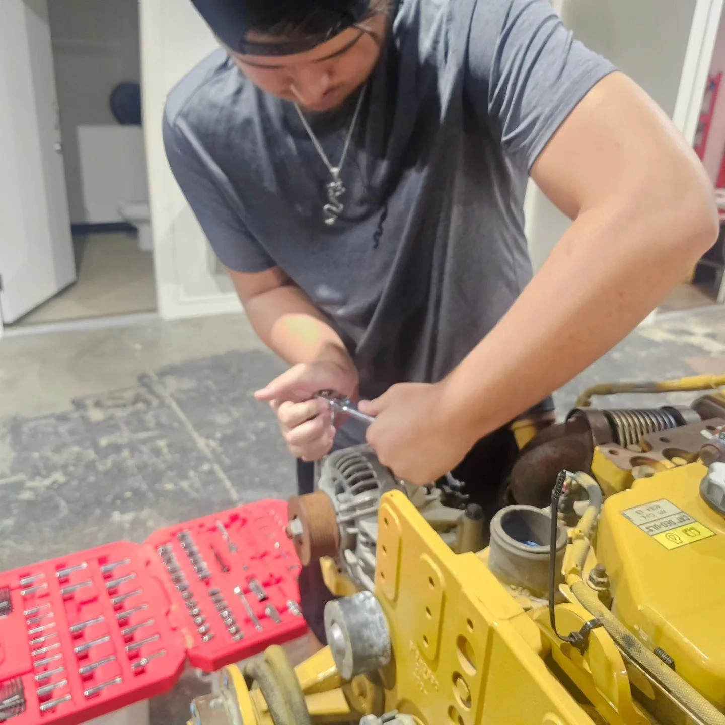 Person wearing a gray t-shirt and a bandana working with tools on a yellow engine, with a red toolbox nearby.