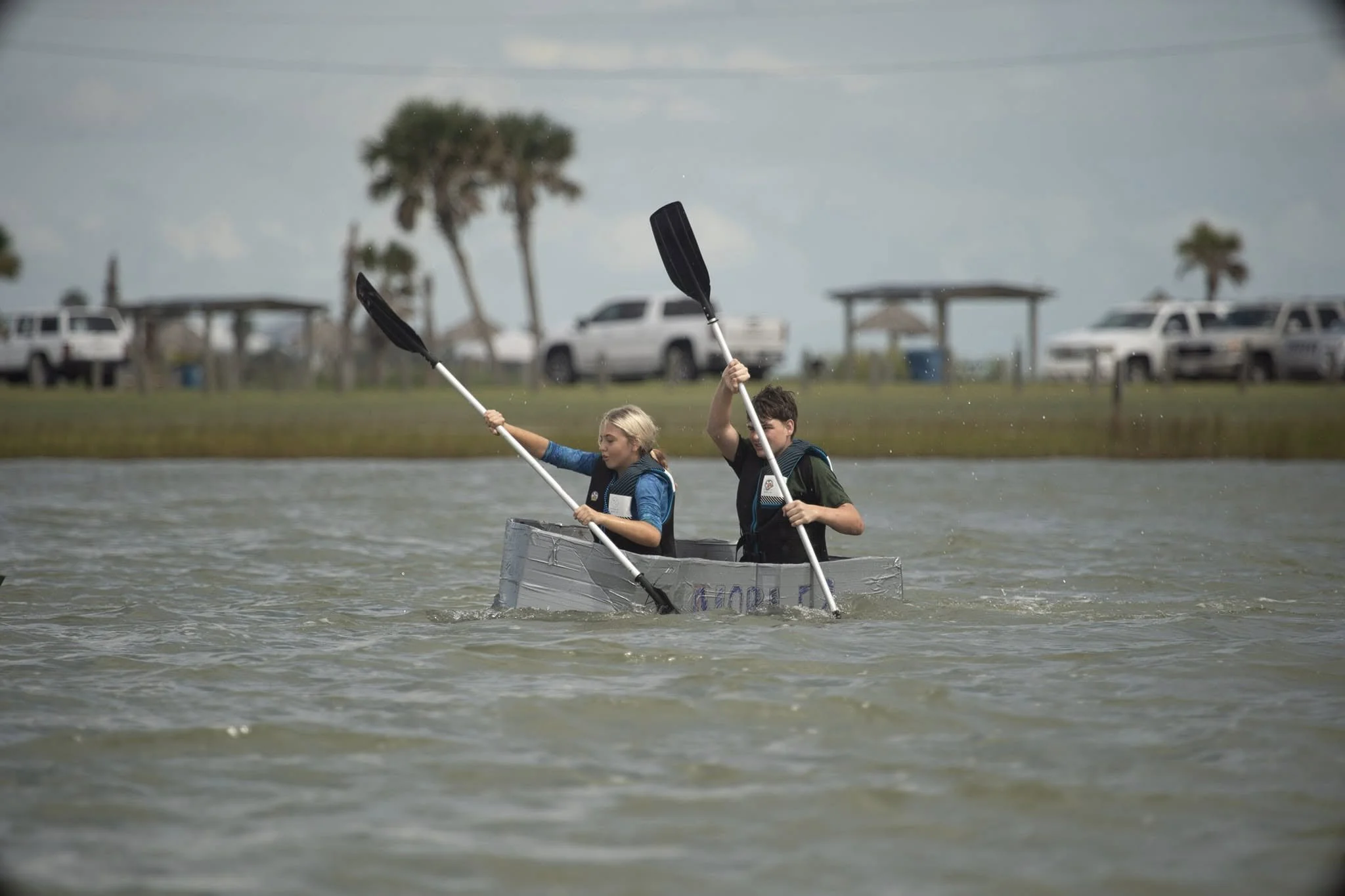 Two children in a handmade boat on a body of water, paddling with oars, with a park-like background featuring trees, parked cars, and small structures.