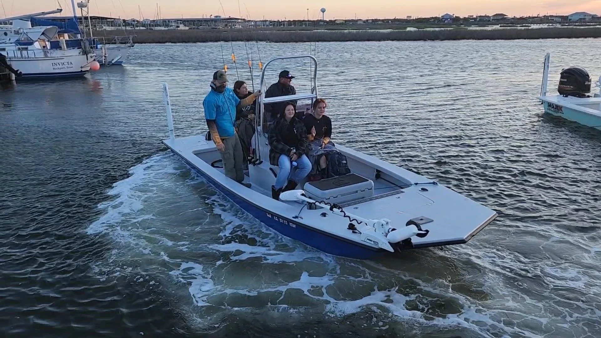 Maritime club. A group of five people on a small white and blue motorboat sailing in a harbor with other boats and shoreline houses in the background during sunset.